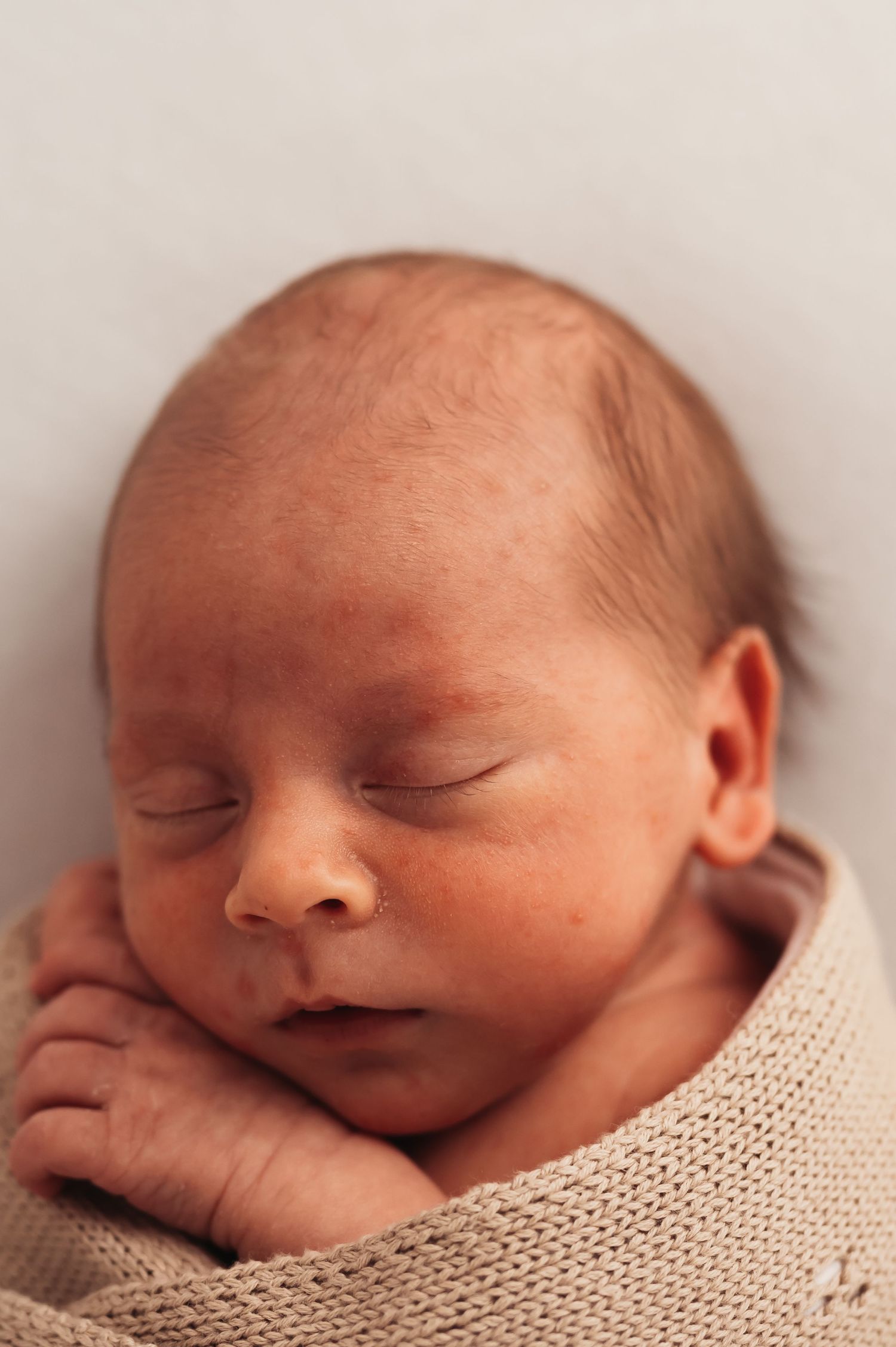 Sleeping infant swaddled in tan knit blanket against neutral background.