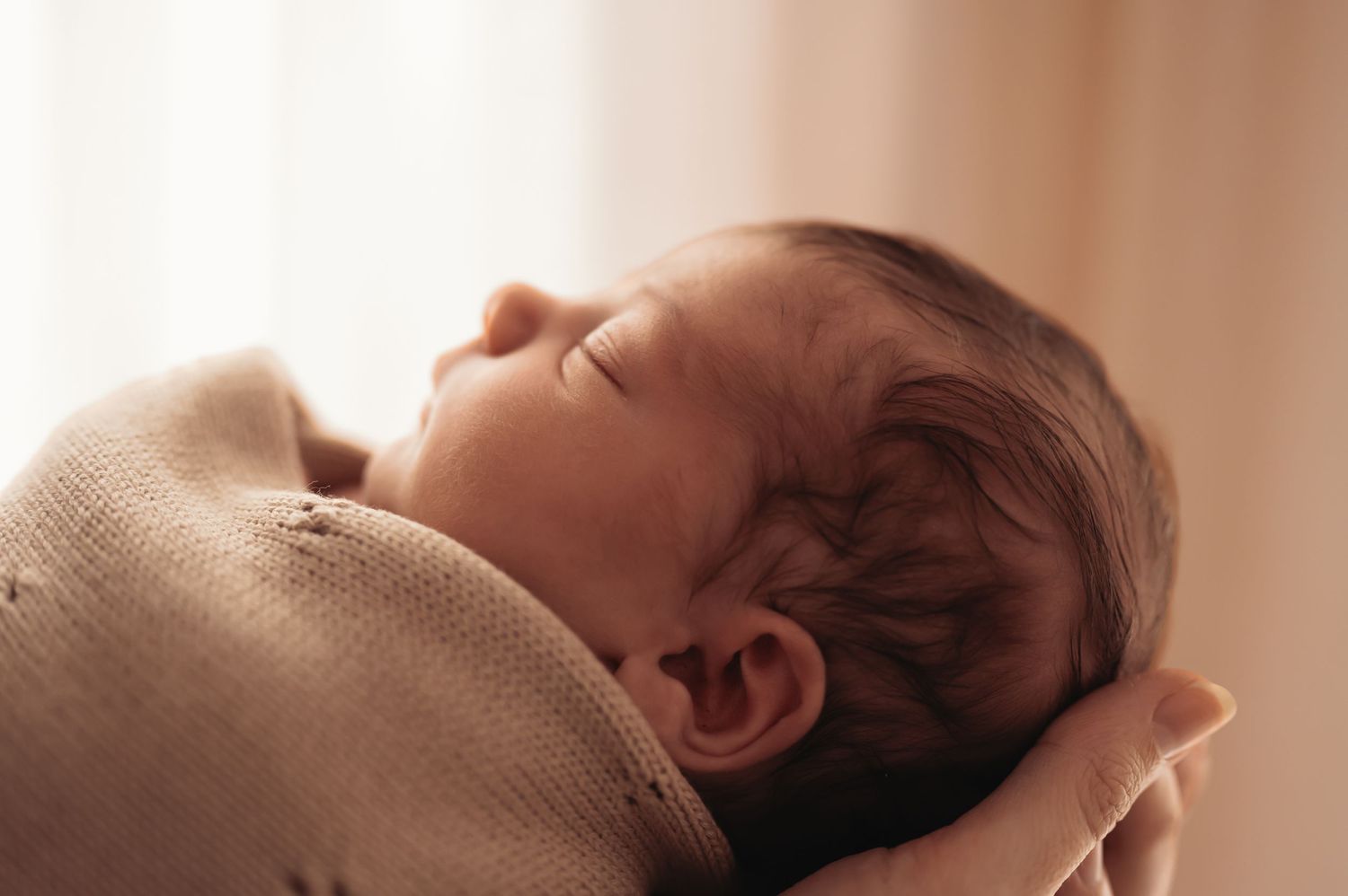 Side view of sleeping infant wrapped in cozy beige blanket in warm lighting.