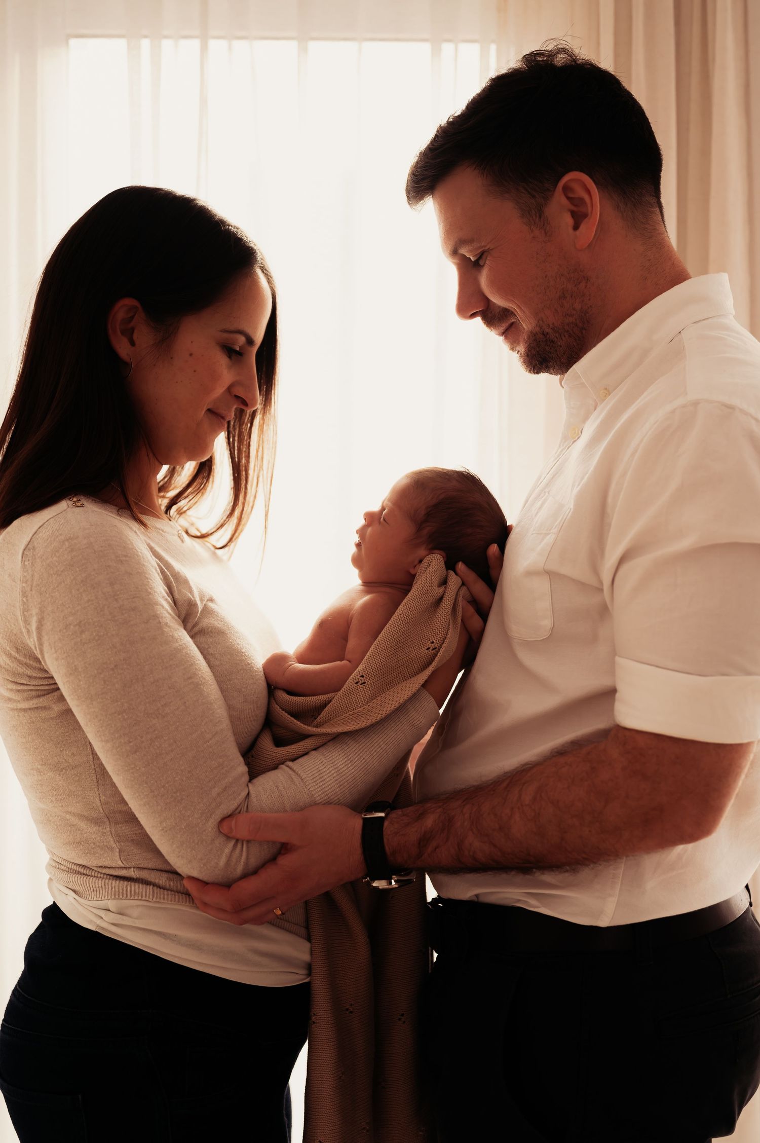 Parents sharing intimate moment with their swaddled newborn by window with soft lighting.