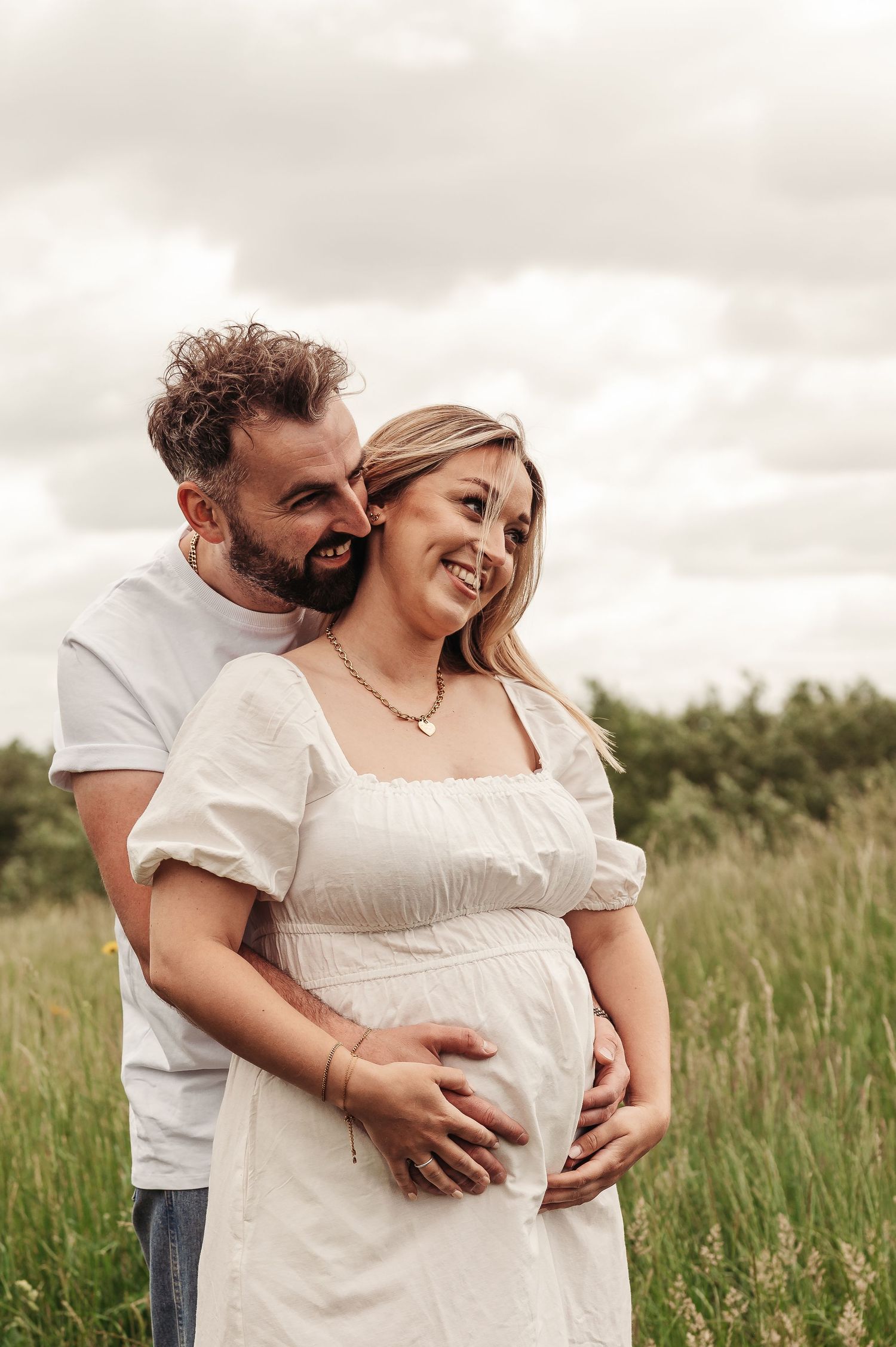 A couple embraces in a sunny meadow with tall grass, sharing a tender moment as they celebrate an expected arrival.