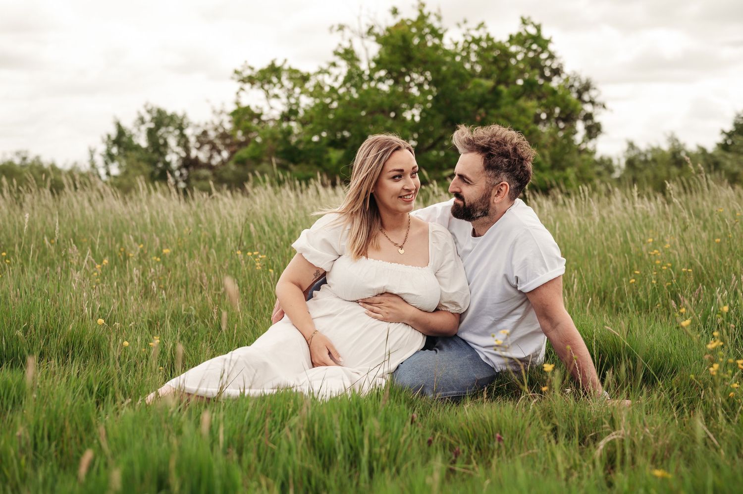 Expecting couple sits together in a serene meadow with tall grass on a cloudy summer day.