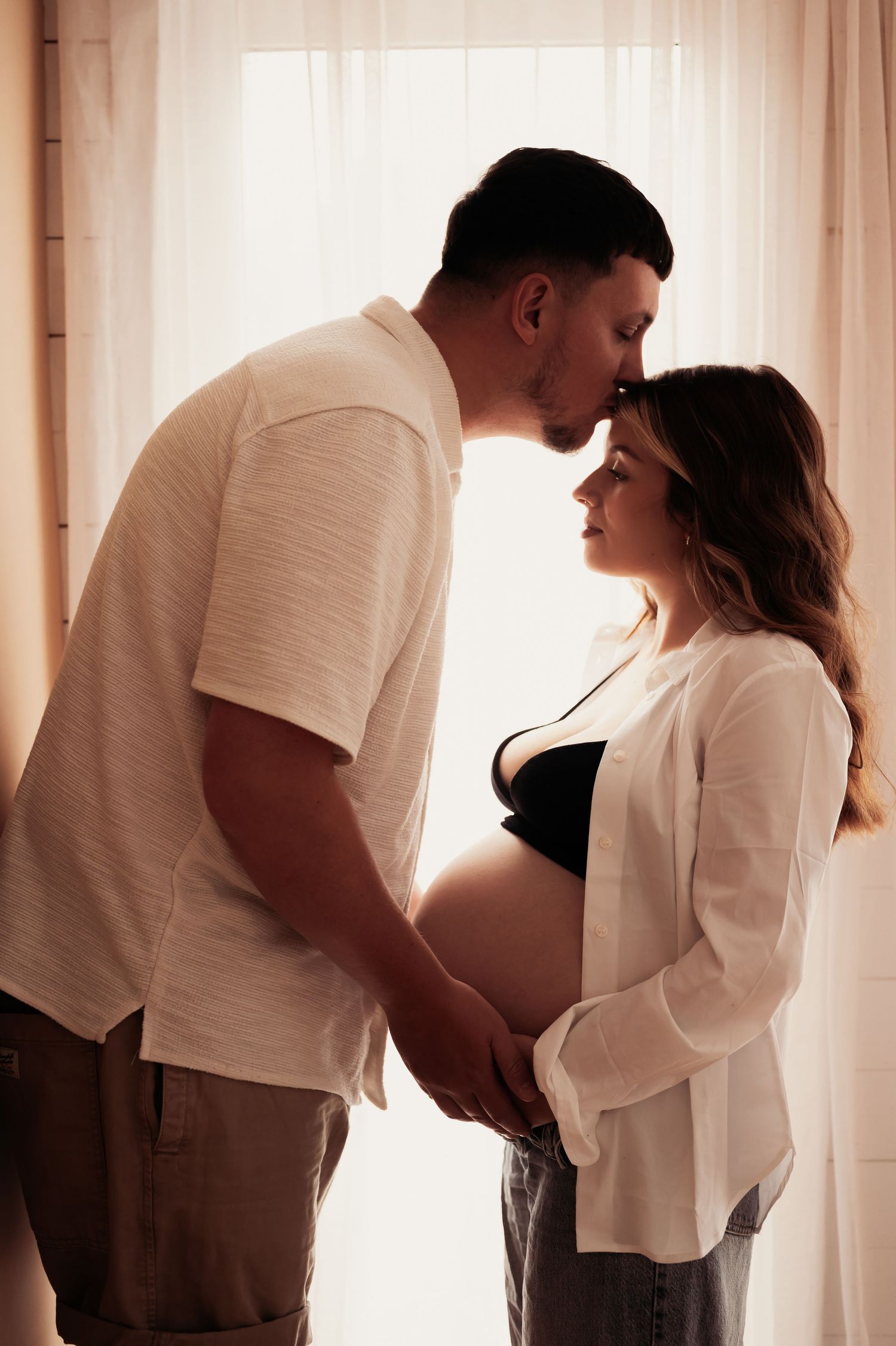 Couple sharing tender moment, pregnant woman holding baby bump, backlit by sheer curtains in soft natural light.