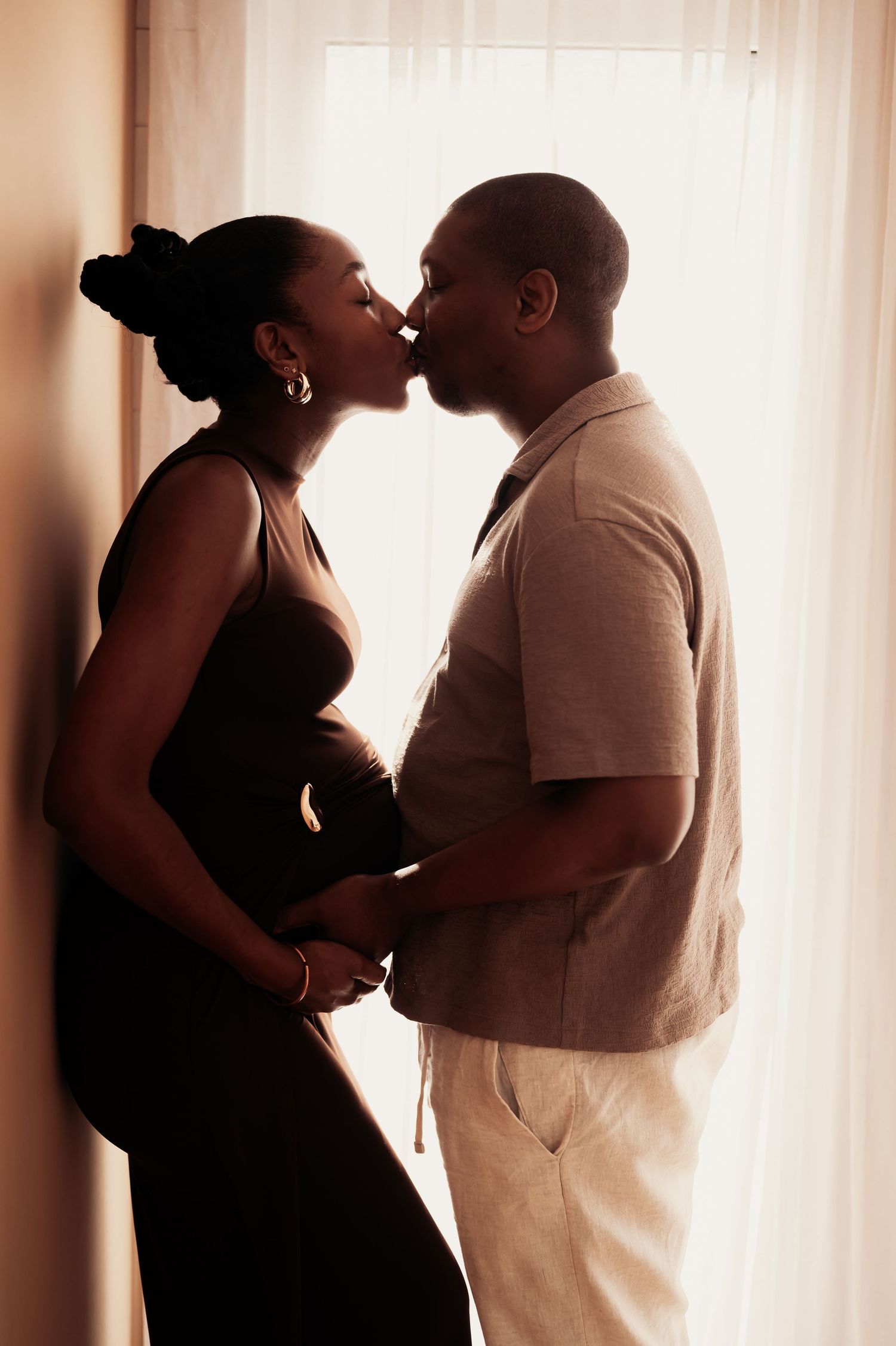 A pregnant couple shares a tender kiss while holding her baby bump, silhouetted against a bright window.