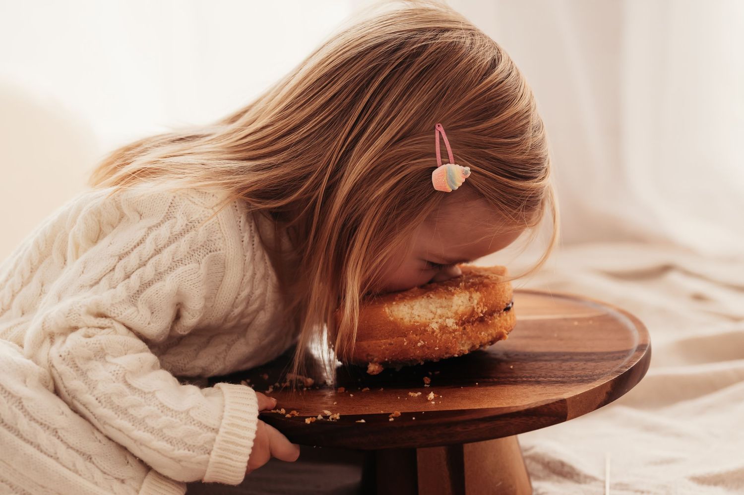 Child in cream sweater leaning over wooden cake stand to eat cake.