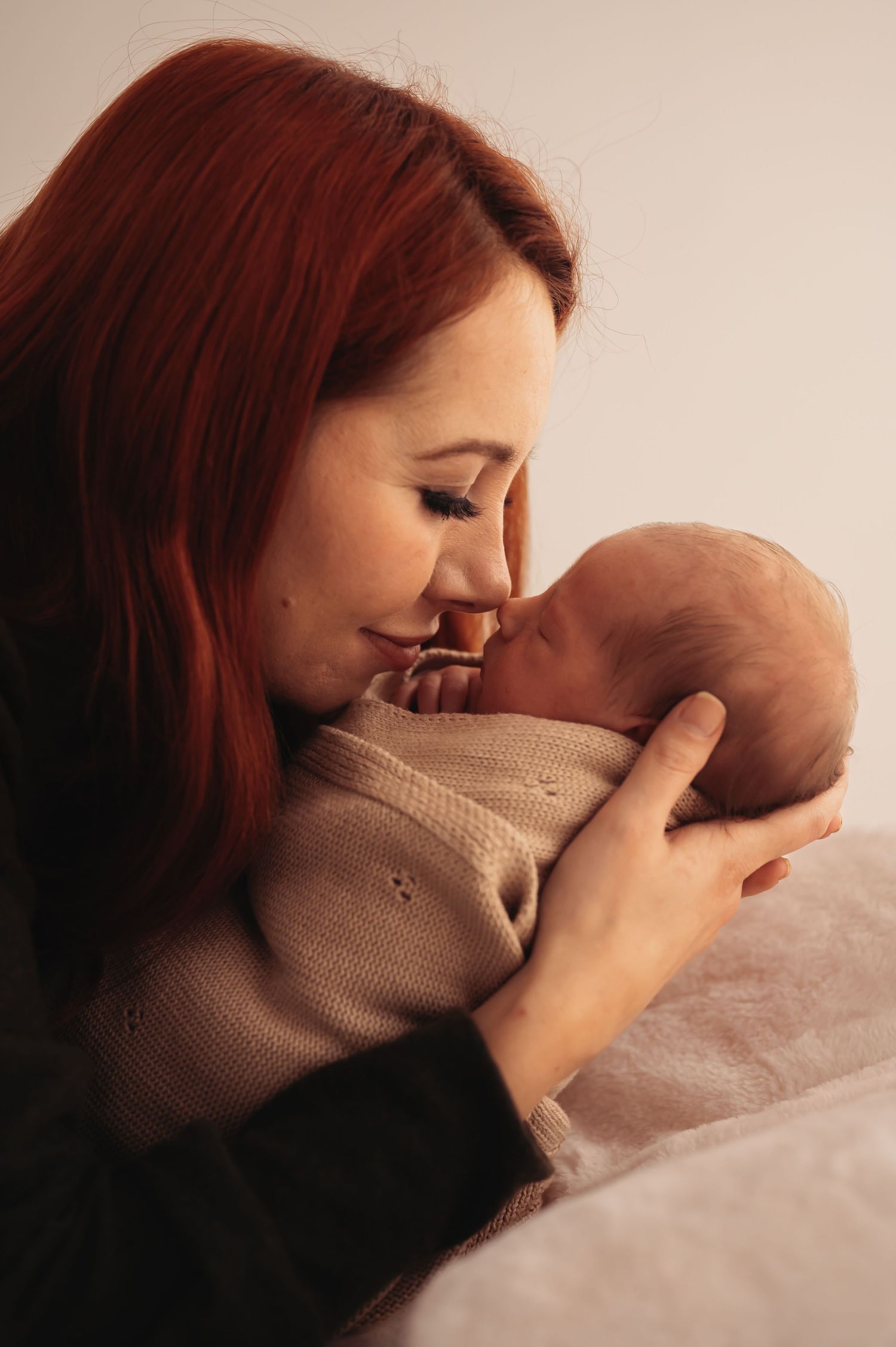 A woman with red hair tenderly cuddles a newborn baby wrapped in a beige knit blanket.