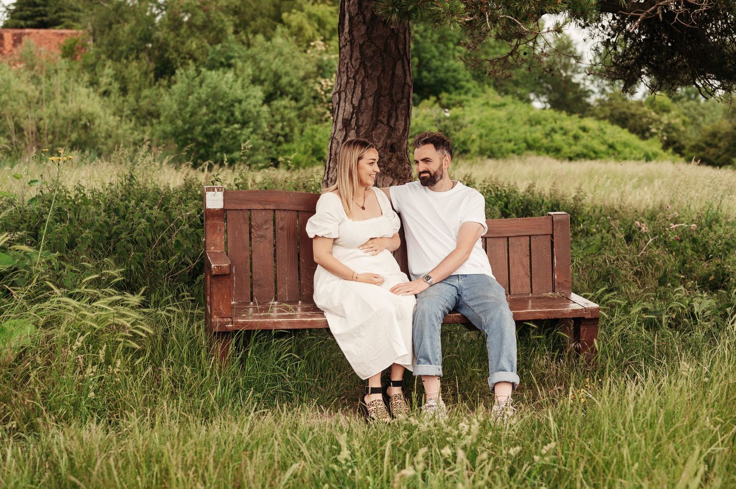 A couple sits together on a rustic wooden bench surrounded by tall grass and lush greenery on a sunny day.