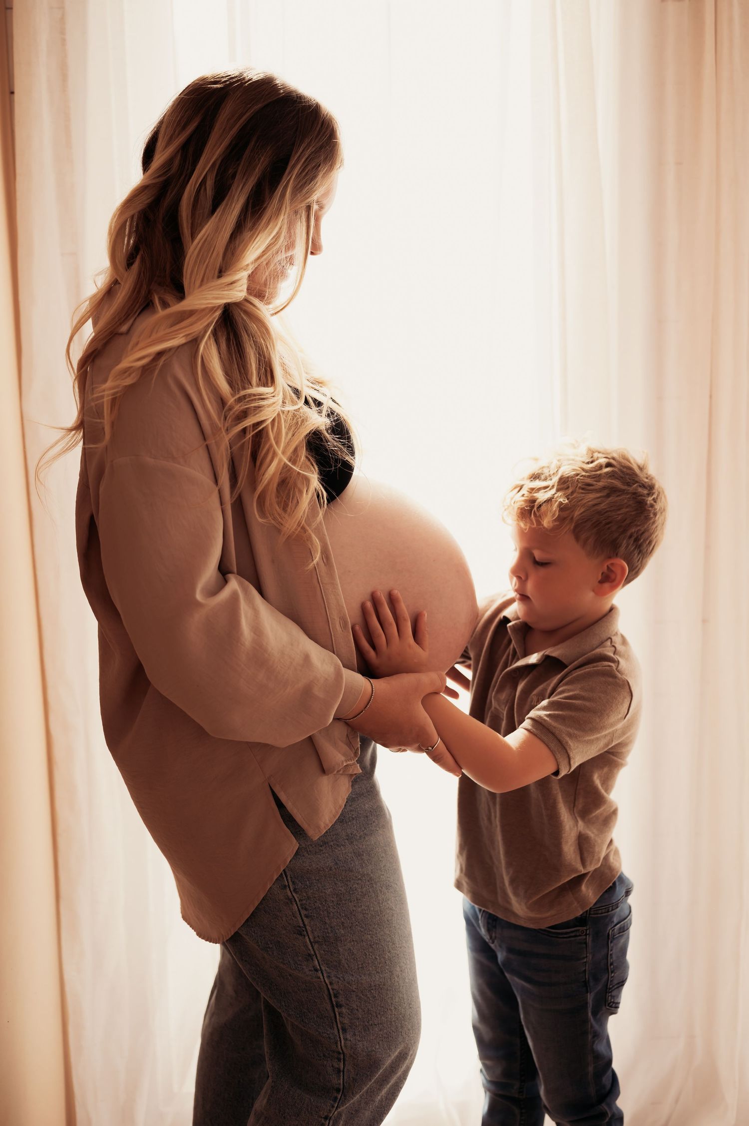 Pregnant woman in beige top with young blond boy touching her baby bump near white curtains.