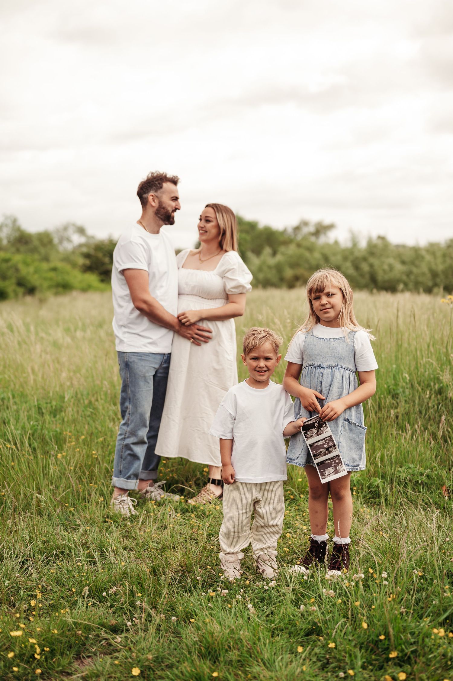 A family wearing white and neutral clothing stands together in a meadow holding ultrasound photos and smiling.