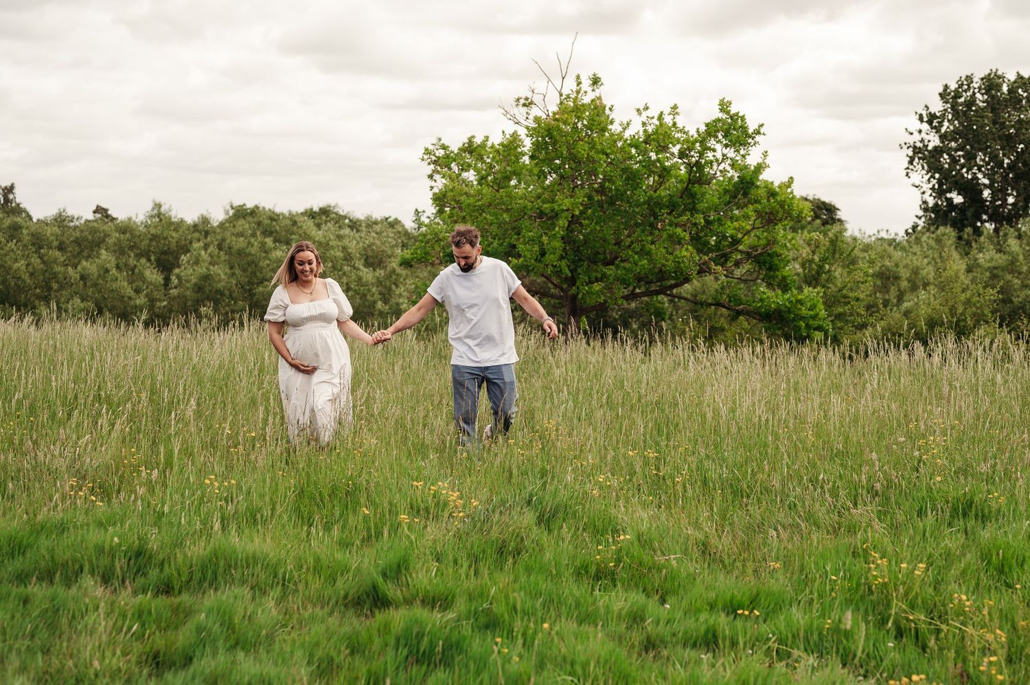 Two people hold hands while walking together through a lush green field on an overcast summer day.