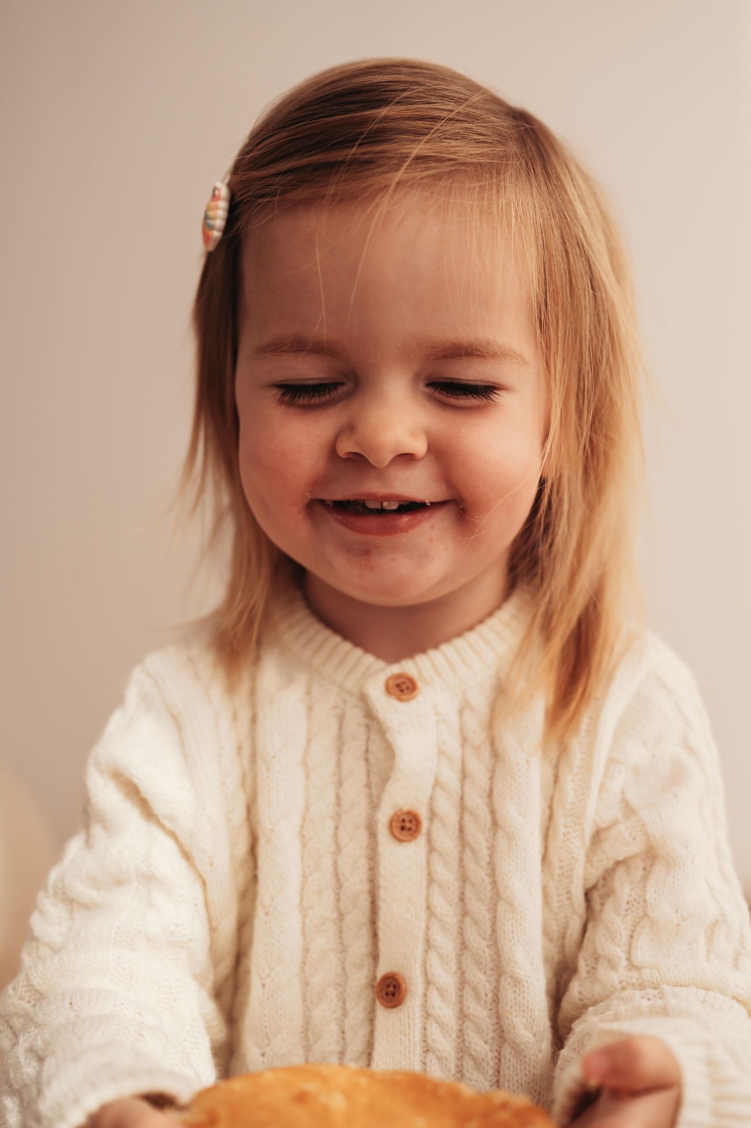 A toddler wearing a cream cable-knit cardigan smiles while holding cake.