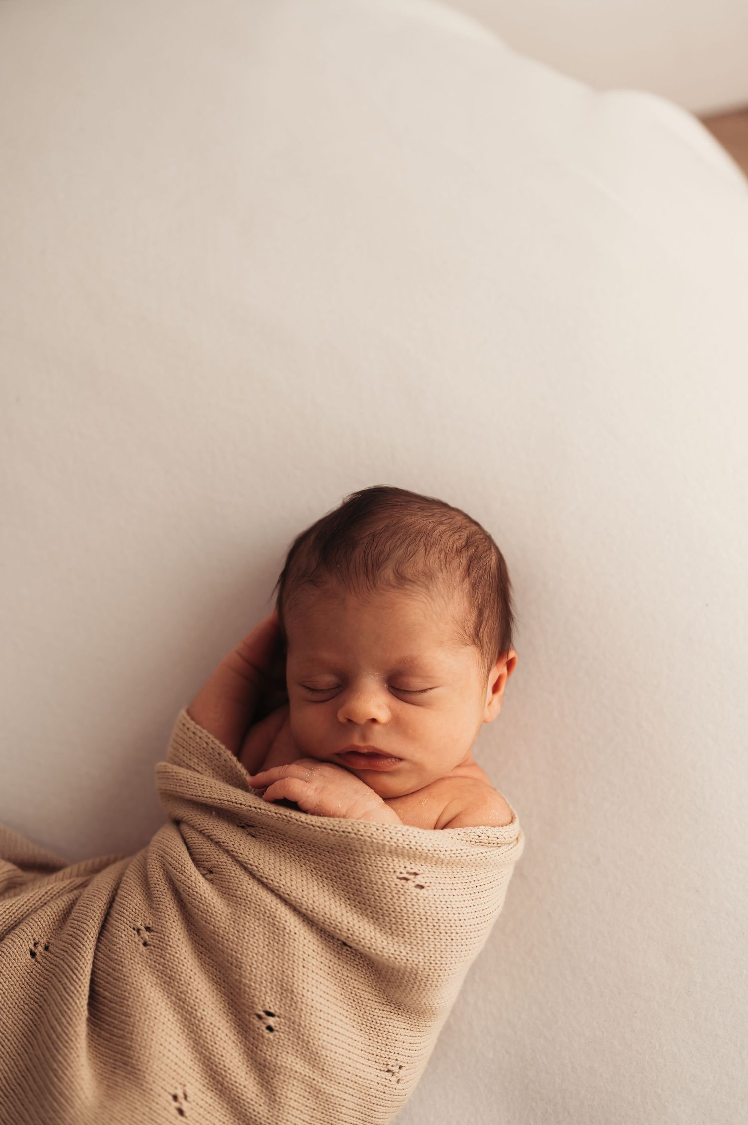 Peaceful sleeping newborn swaddled in beige knit blanket on white surface.