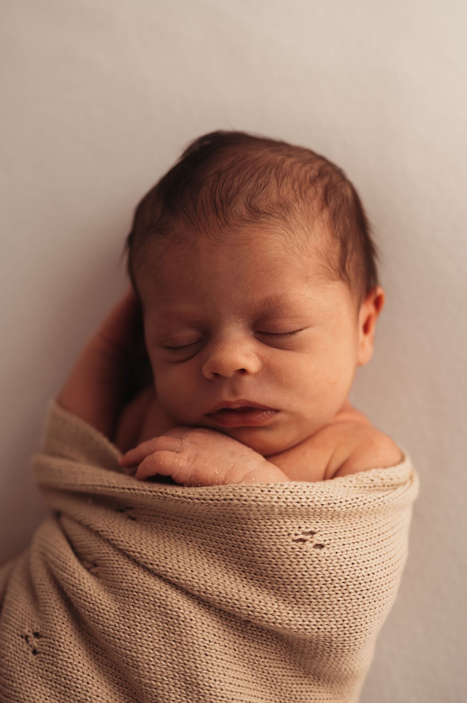 Close-up of sleeping newborn wrapped in textured beige blanket against neutral background.