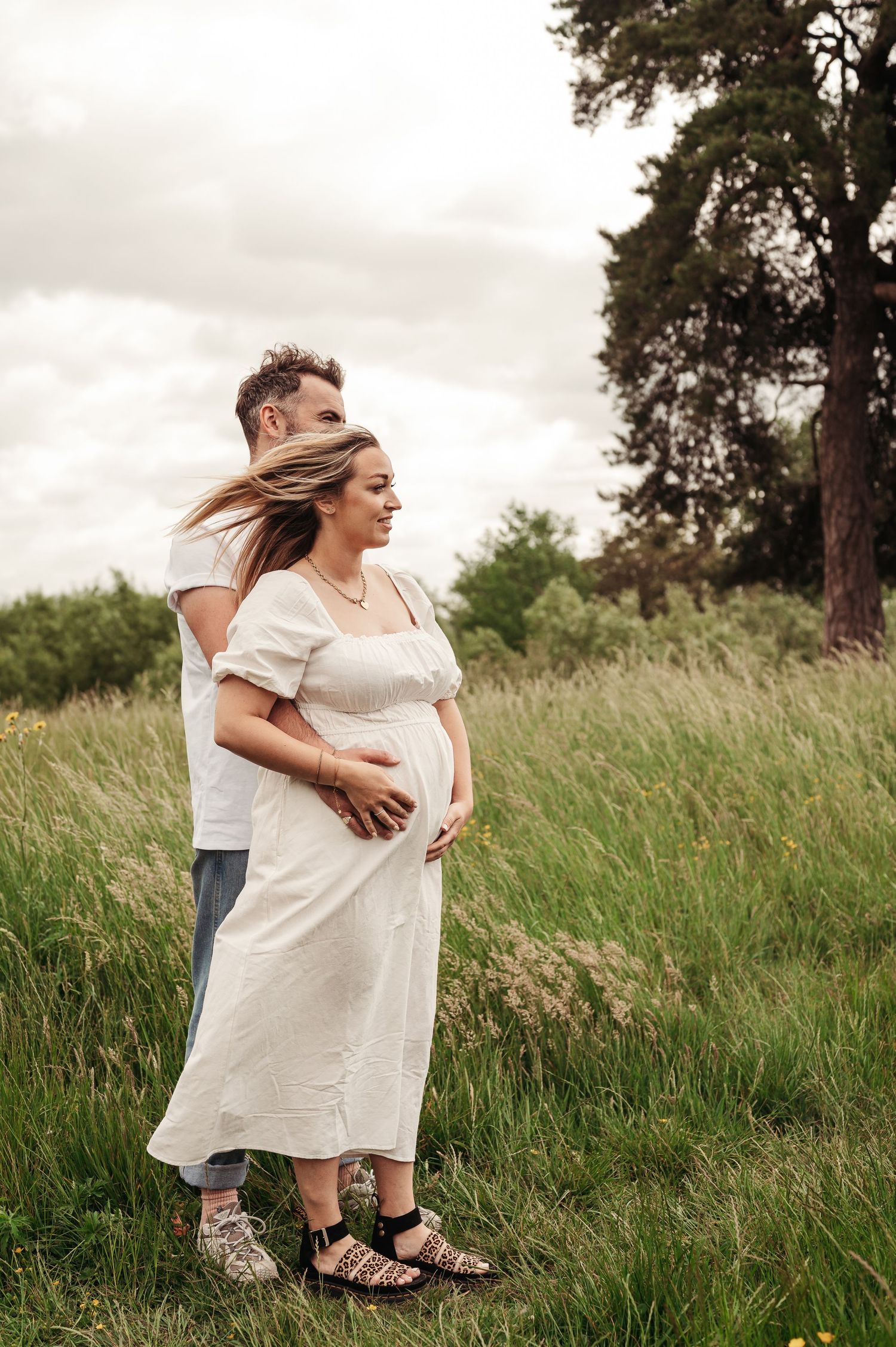 A loving couple shares an intimate moment in a sunlit meadow, with the expectant mother wearing a flowing white dress.