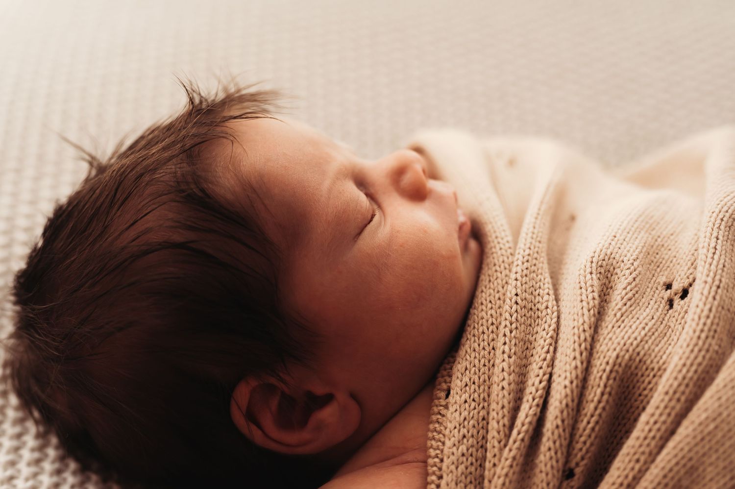 Peaceful newborn baby sleeping wrapped in a soft beige knit blanket with dark hair visible.