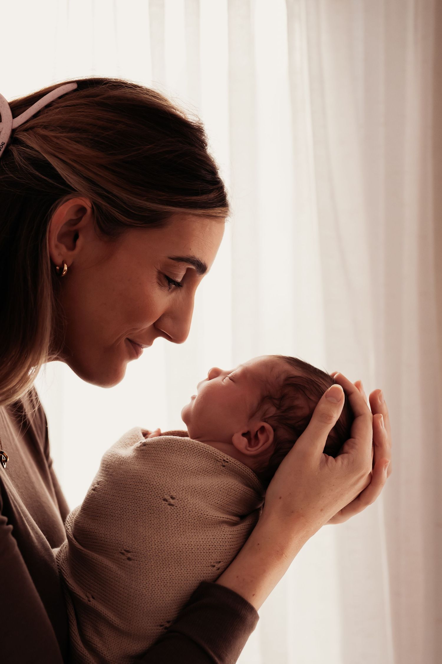 A mother tenderly holds her sleeping newborn baby near a softly lit window, smiling down with love.