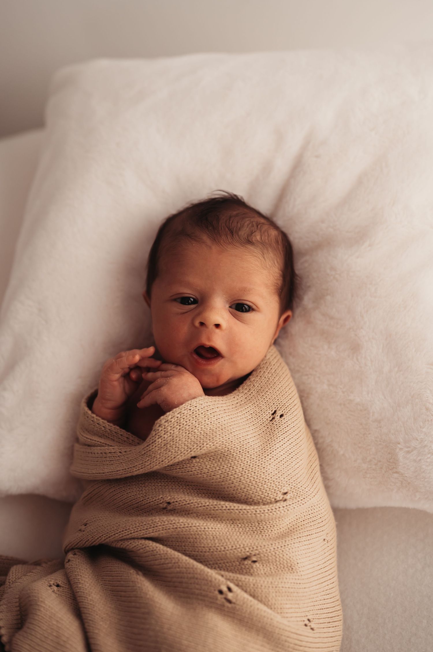 Alert newborn baby swaddled in beige blanket looking at camera on white pillow.