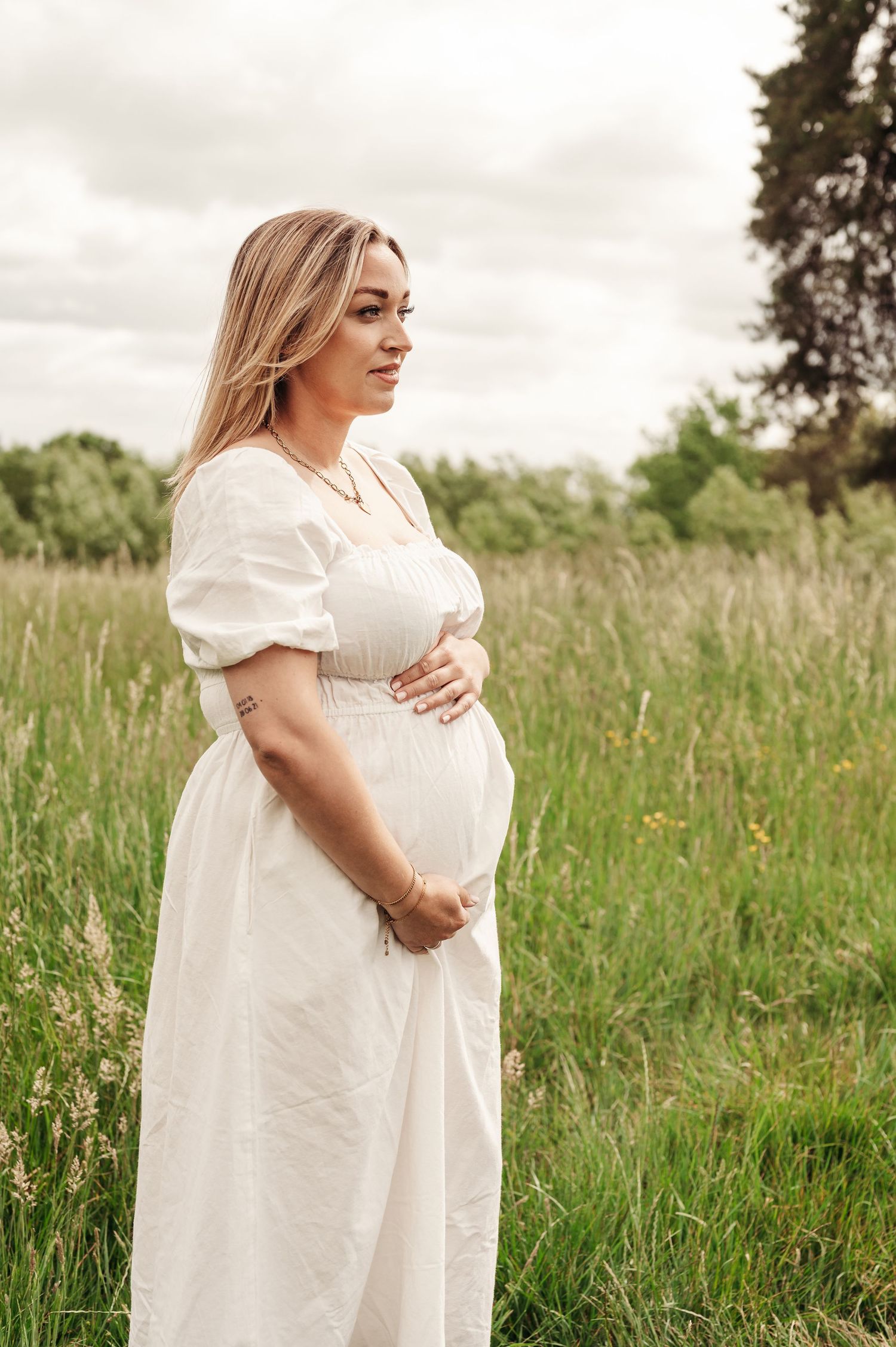 Person in flowing white dress stands thoughtfully in a serene meadow with tall grass and wildflowers.
