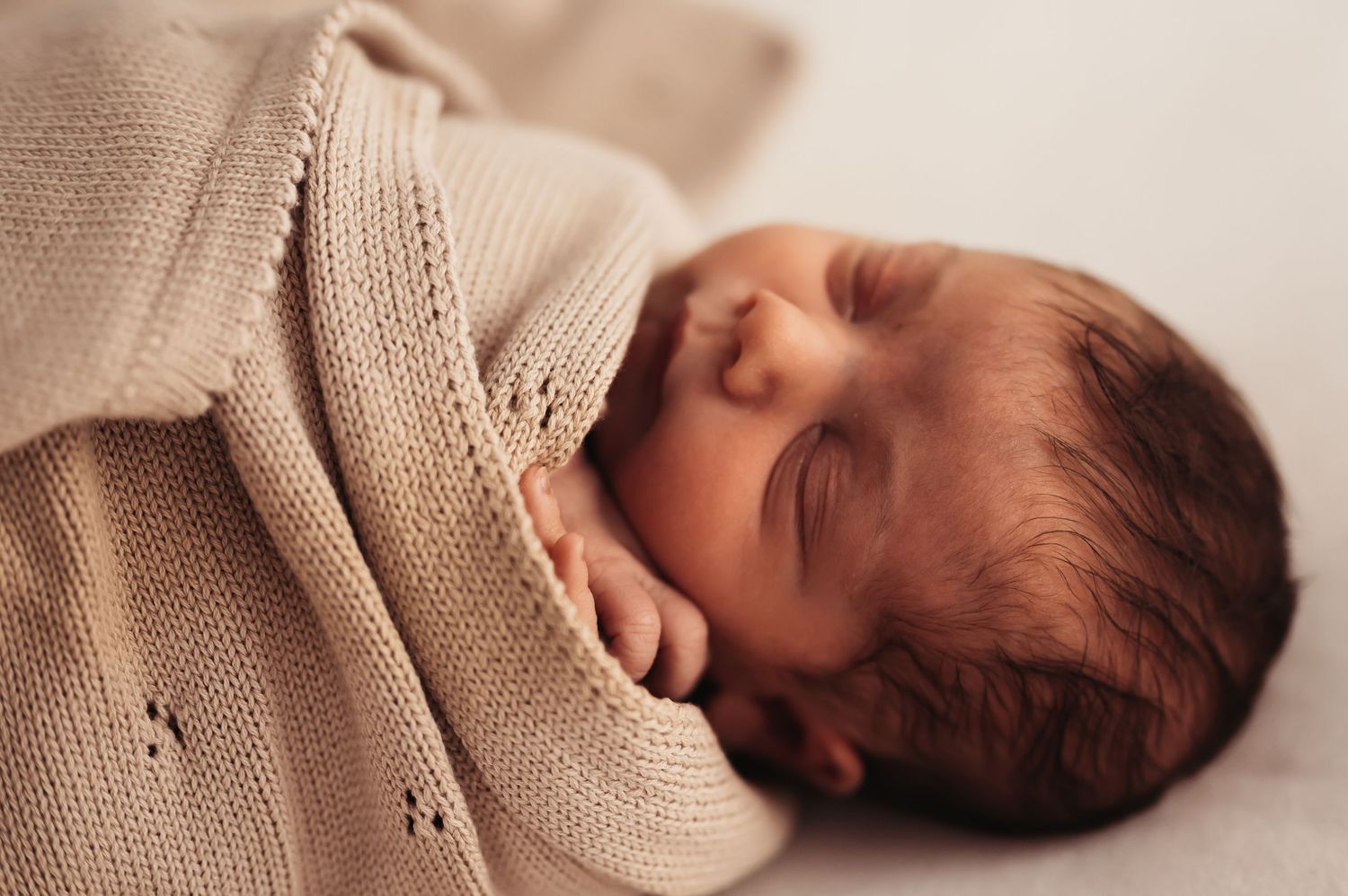 Peaceful newborn baby swaddled in a soft beige knit blanket, sleeping soundly with tiny hands visible.