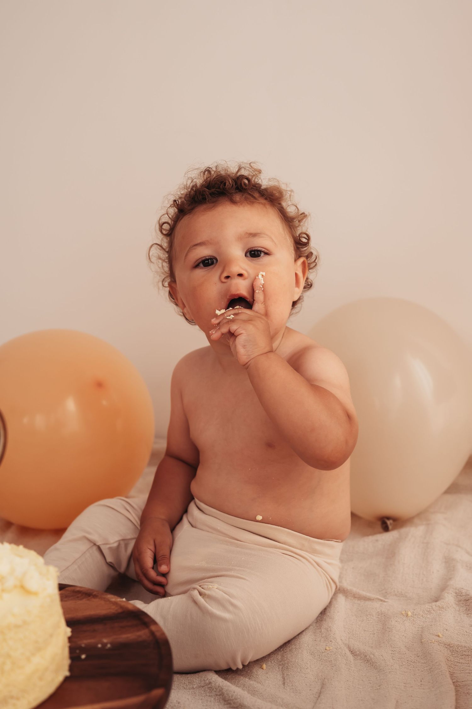 Toddler enjoying cake smash photo session with orange and white balloons in background.