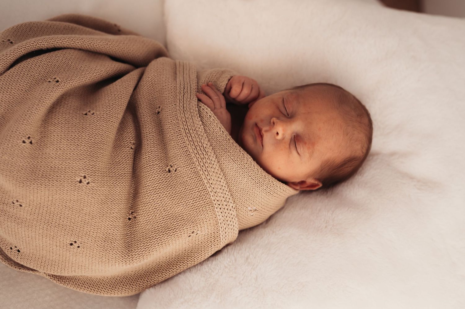 Baby sleeping soundly wrapped in beige patterned knit blanket.