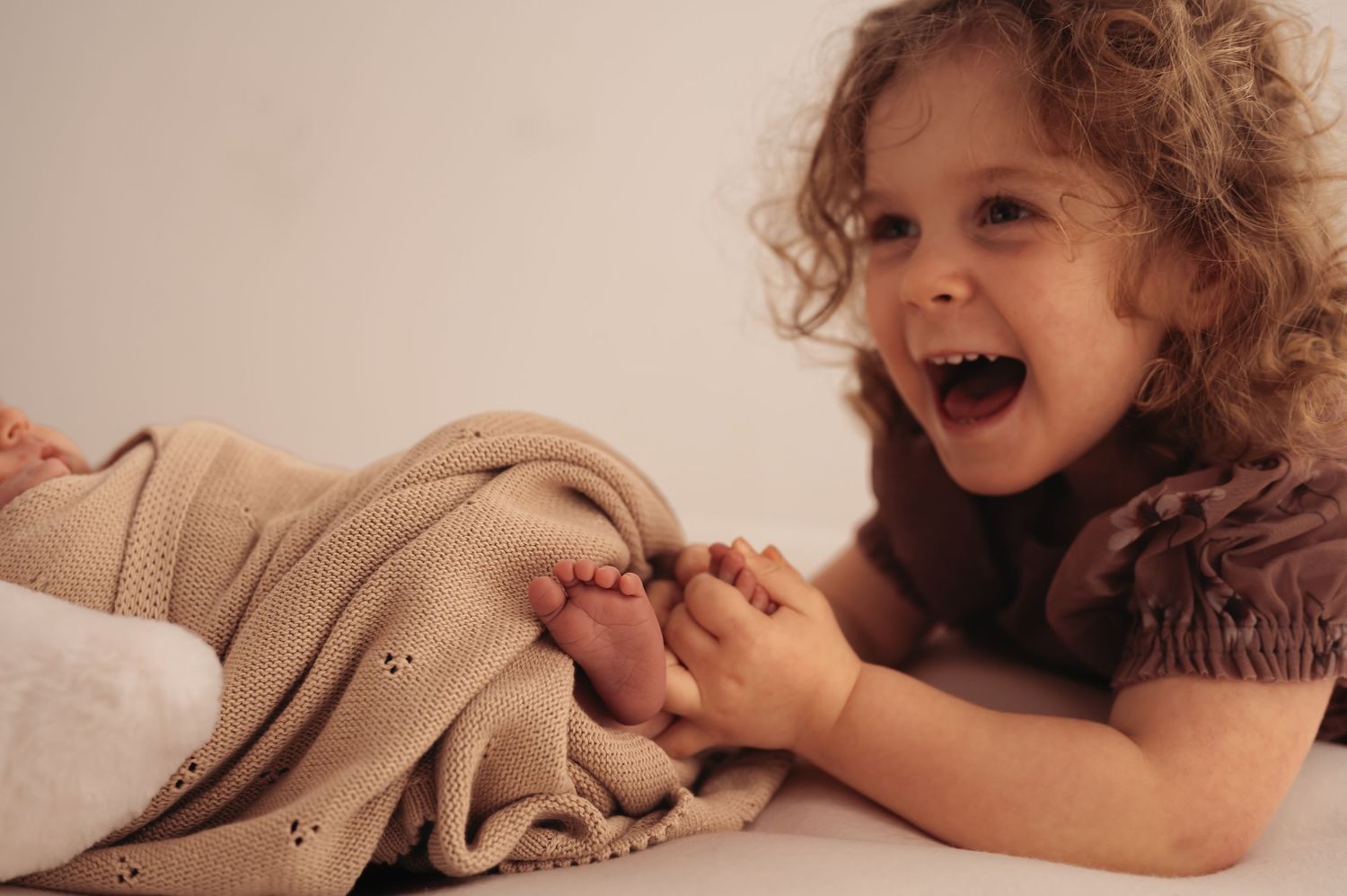 A young child with curly hair laughs joyfully while playing with a beige knit blanket.