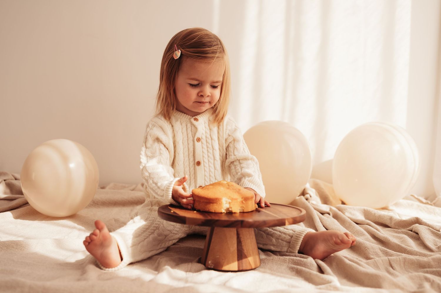 Child in white knit outfit sitting with cake stand and balloons on beige fabric.