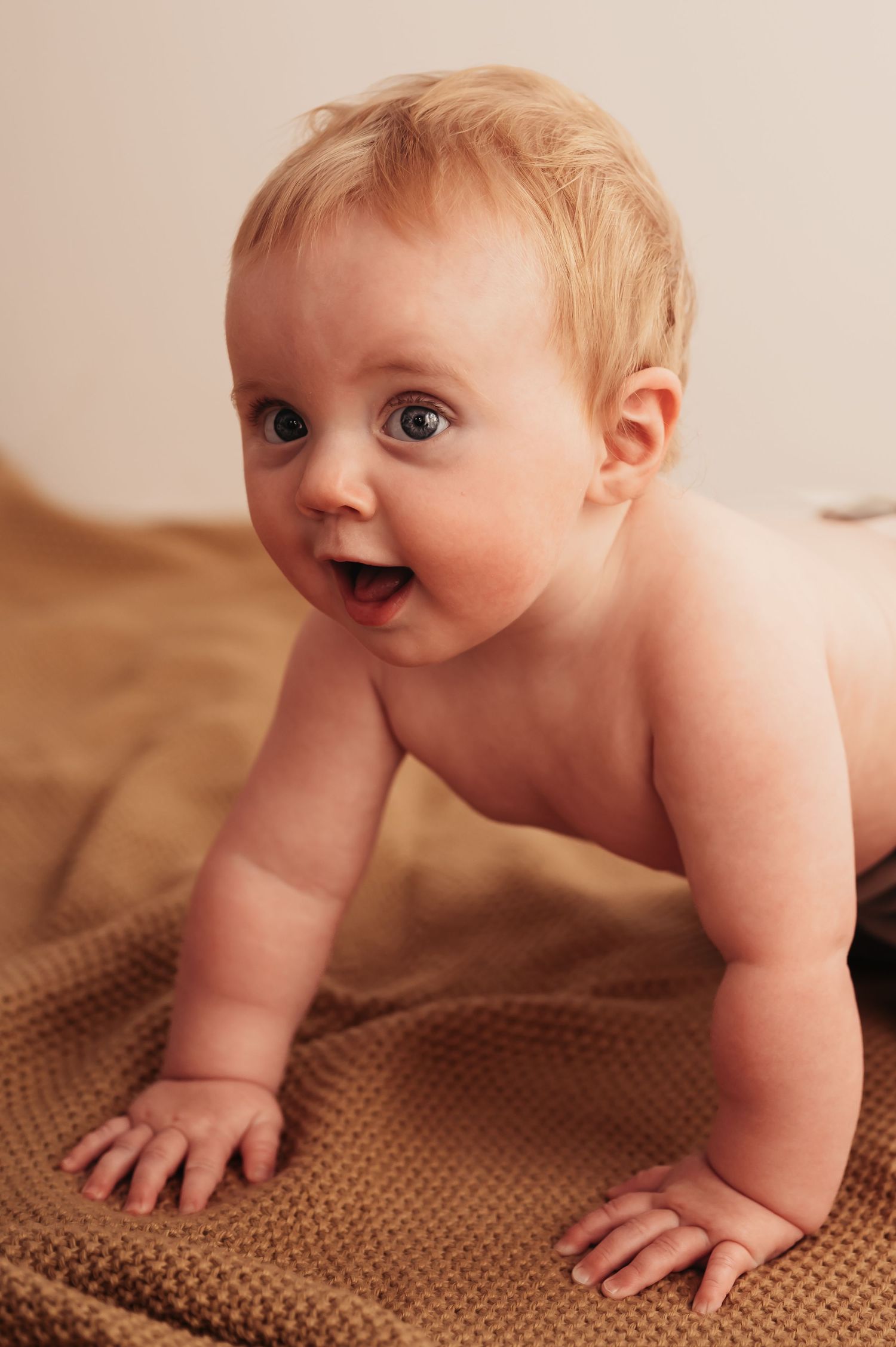 Happy baby with blonde hair crawling on brown knitted blanket with excited expression.