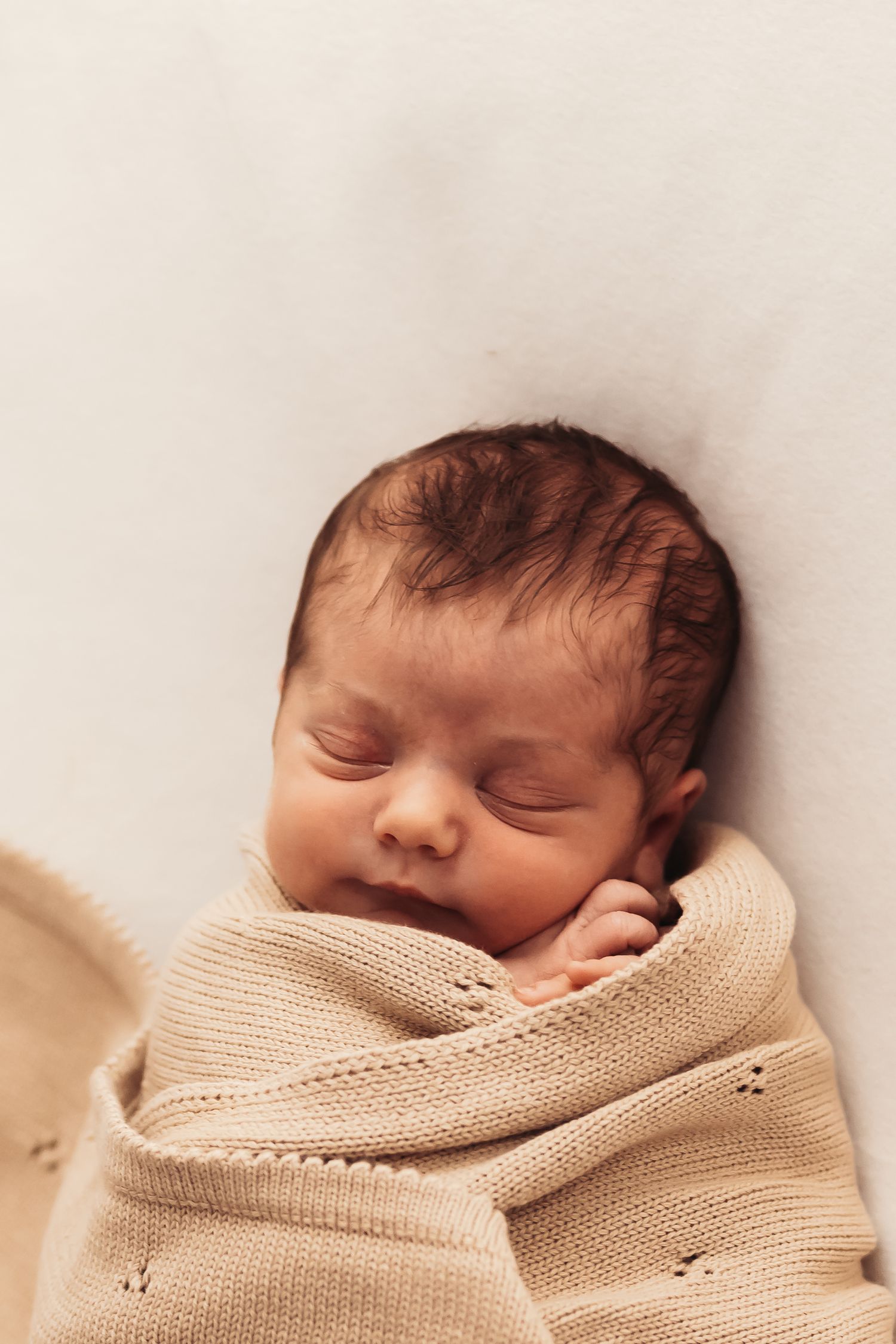 Sleeping newborn swaddled in beige knitted blanket against white background.