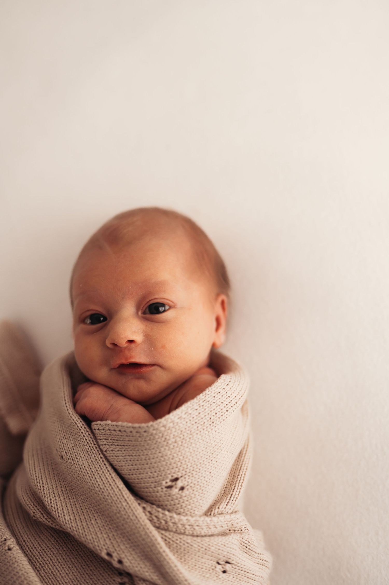 Newborn baby swaddled in beige knit blanket looking at camera.