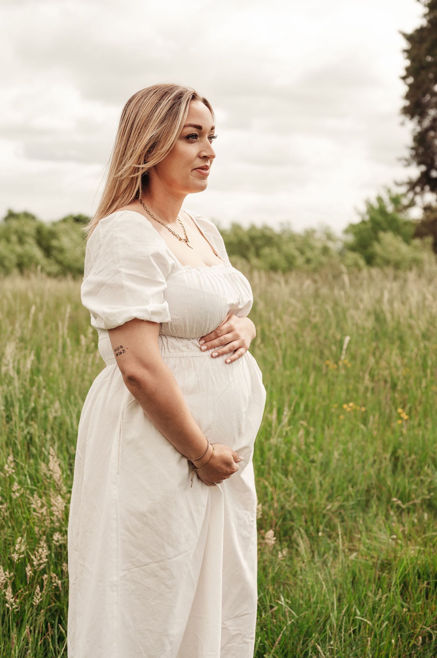 Expectant person wearing a white puffy sleeve dress poses in a peaceful field at sunset.