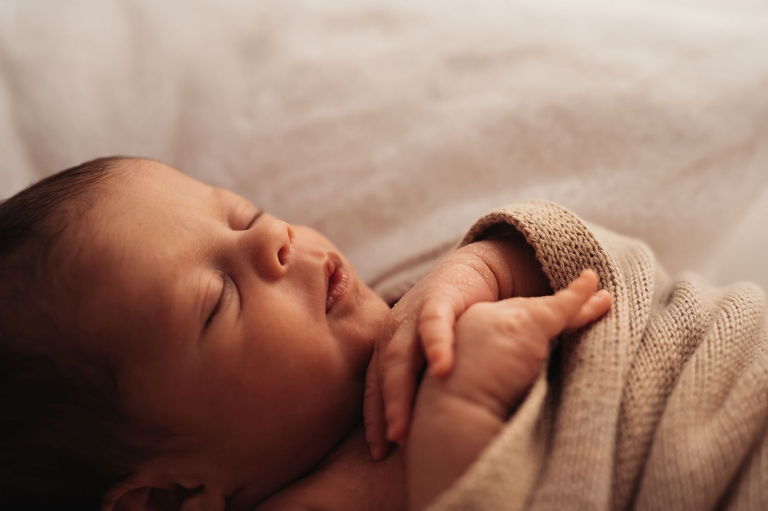 Sleeping baby swaddled in knitted blanket with hands near face on white bedding.