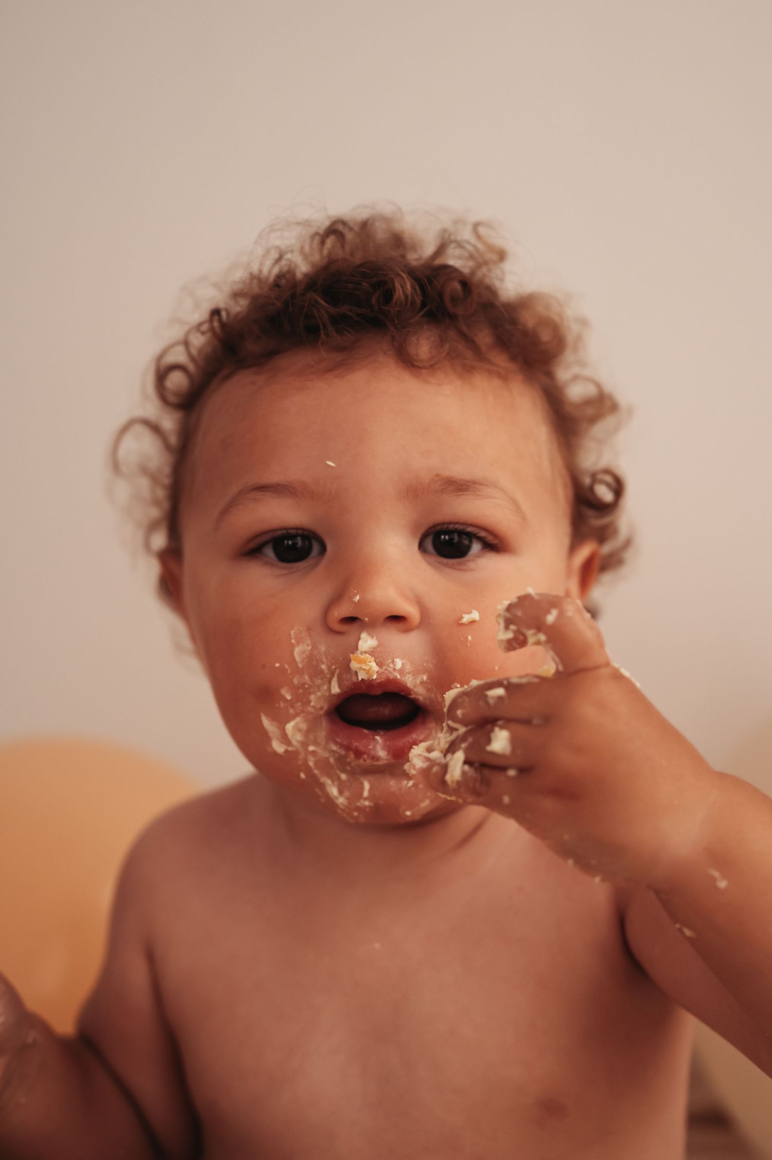 Baby with curly hair touching frosting-covered face during cake smash photography session.