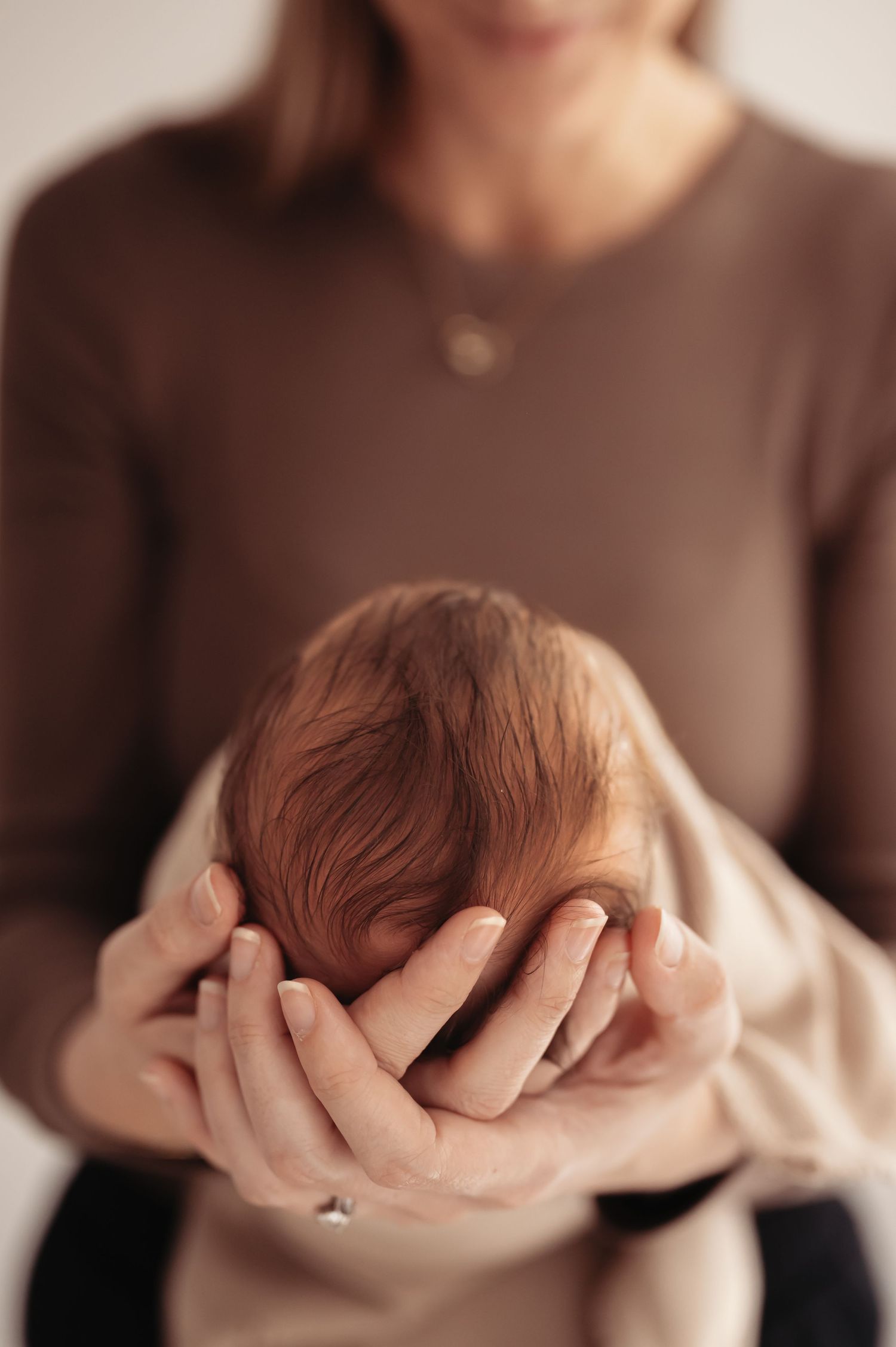 Gentle hands cradle a newborn baby's head wrapped in a soft beige blanket against a warm neutral background.