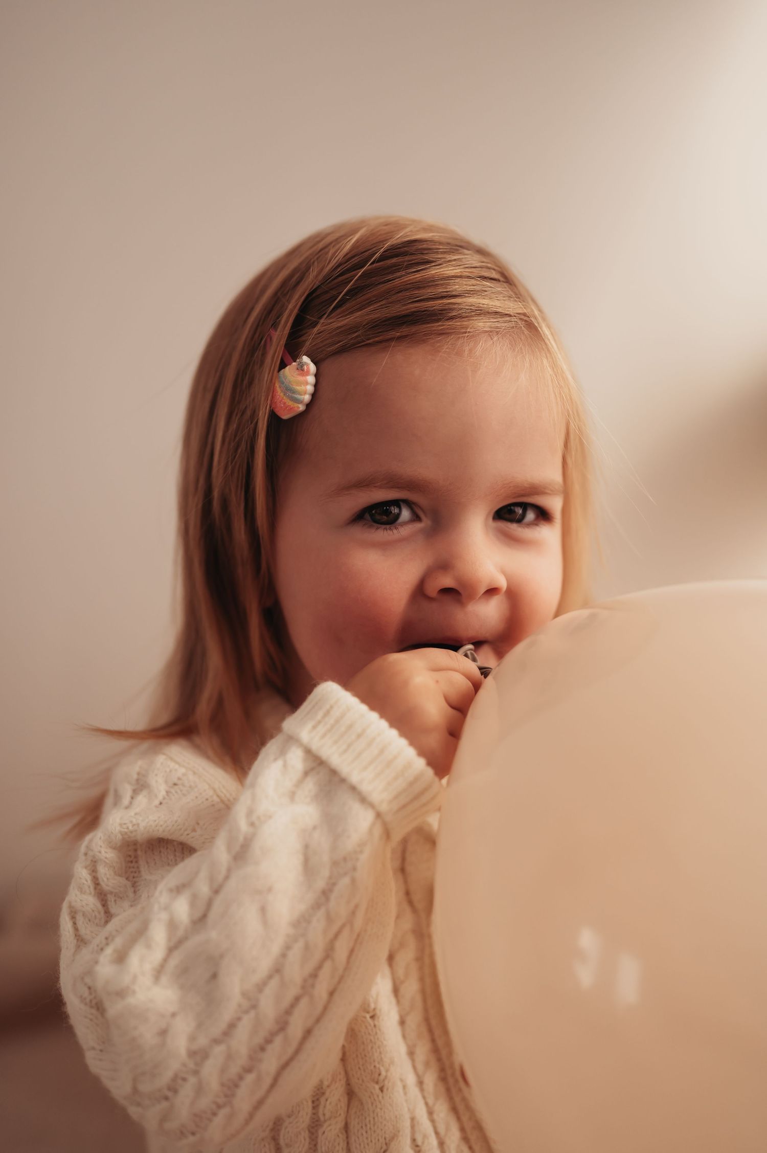 Young child in white sweater smiling shyly while playing with balloon.