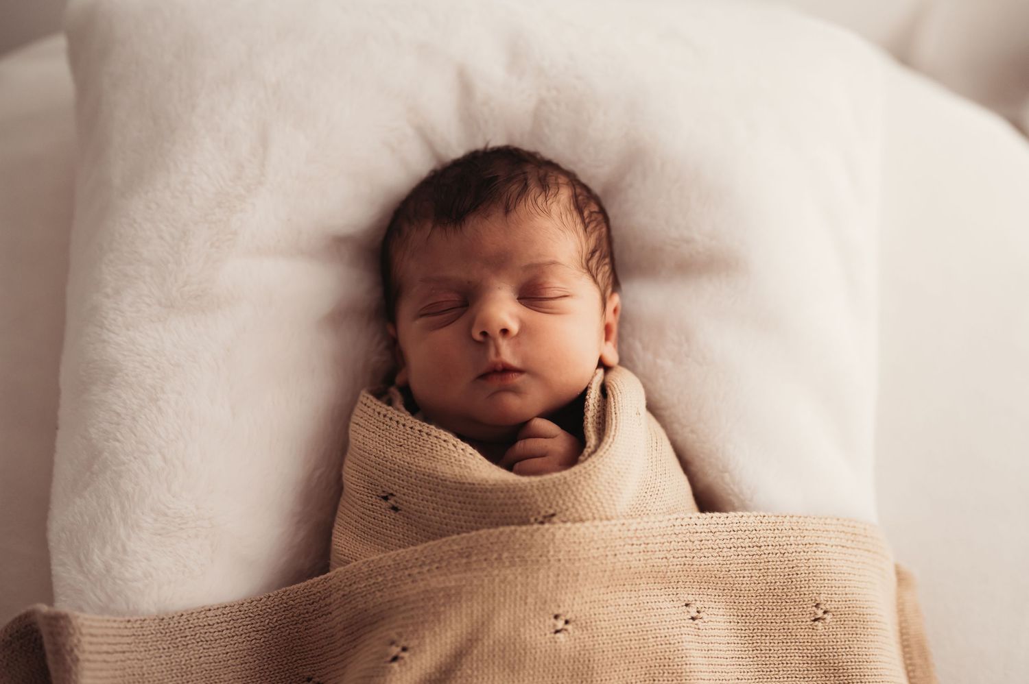 Newborn baby sleeping soundly while swaddled in tan blanket on white pillow.