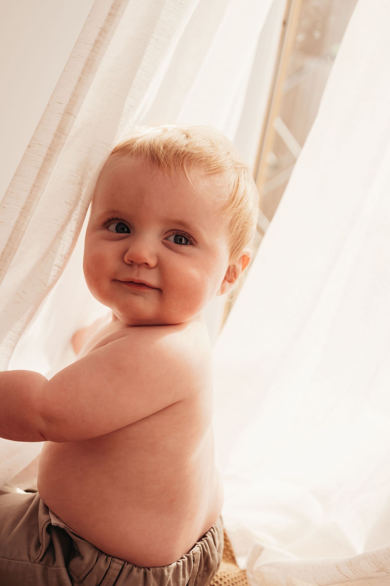 Happy baby standing by sheer curtains in natural window light.