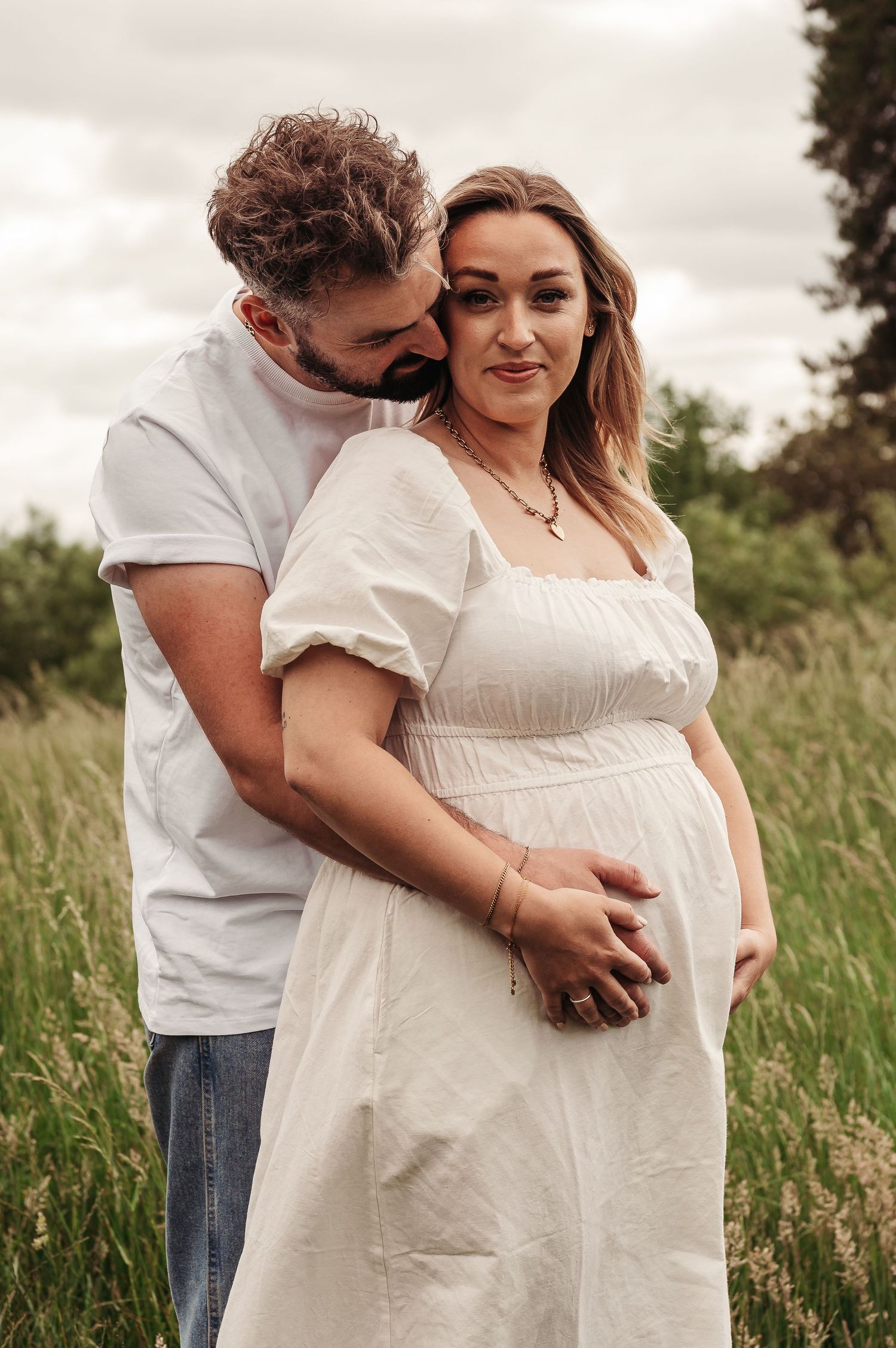 Couple embraces in golden field as they share intimate maternity moment wearing white dress and casual attire.