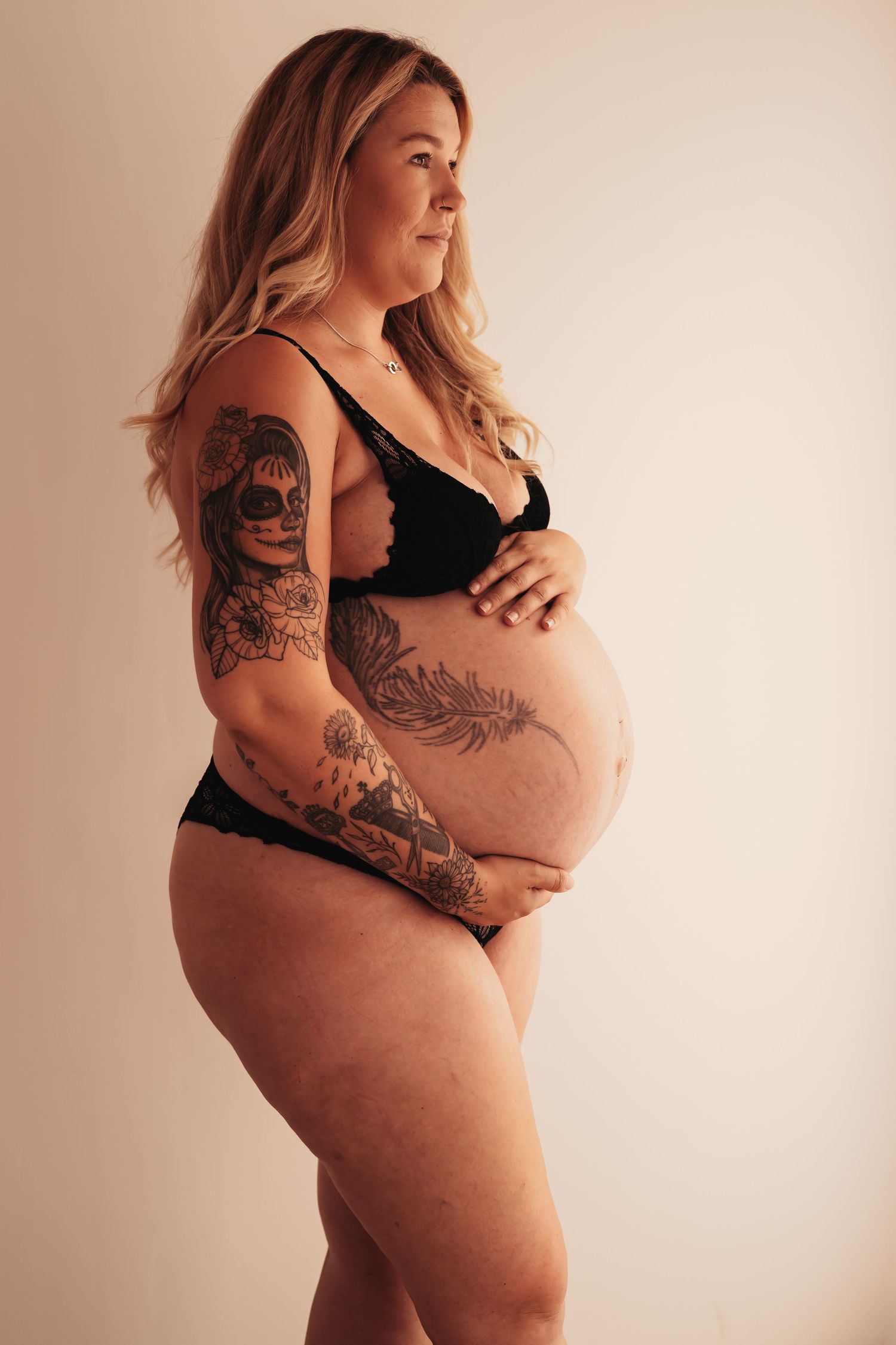 Pregnant woman with tattoos in black lingerie poses against white wall.