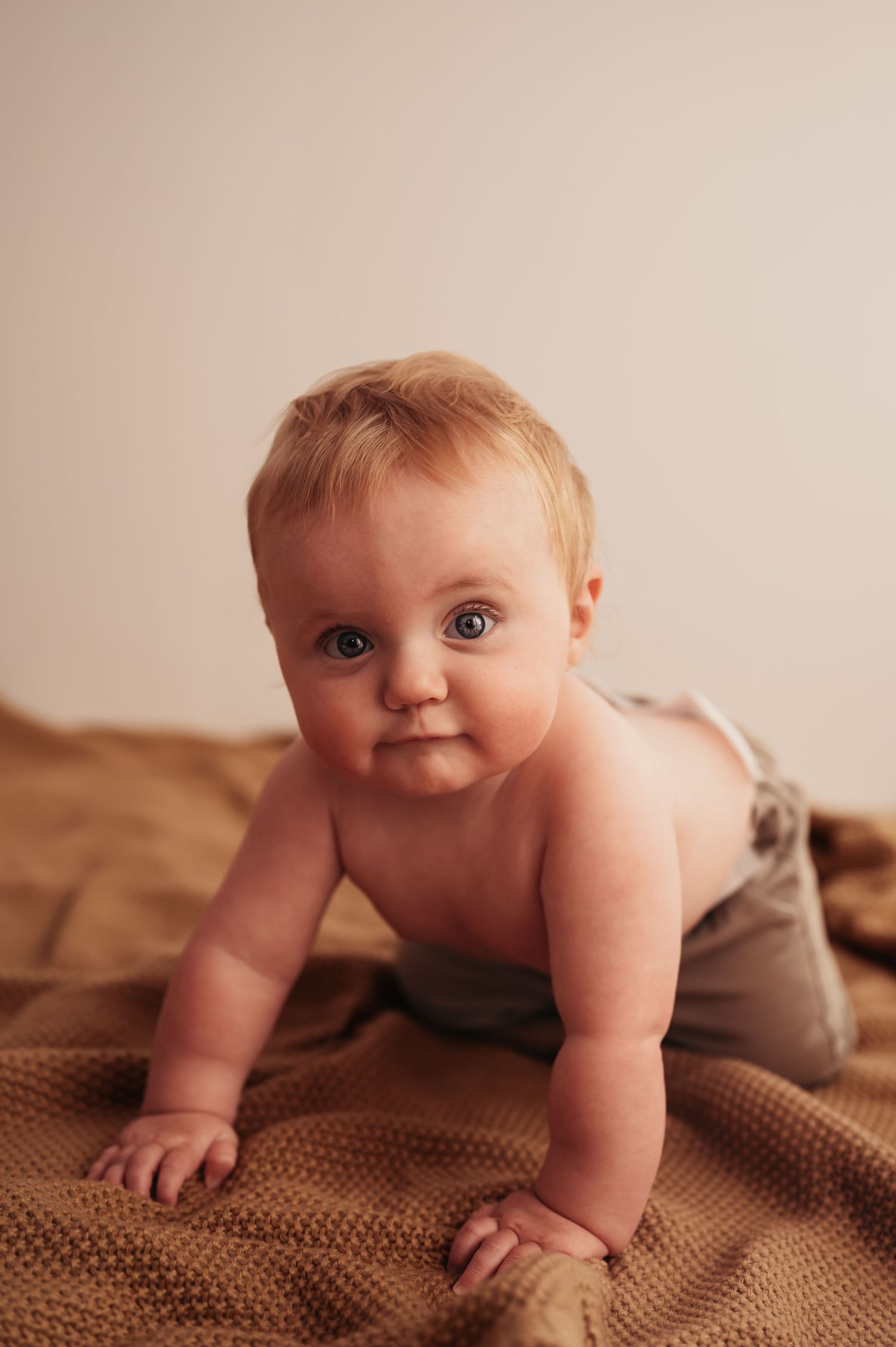 Baby in diaper crawls on brown knit blanket during indoor photo session.