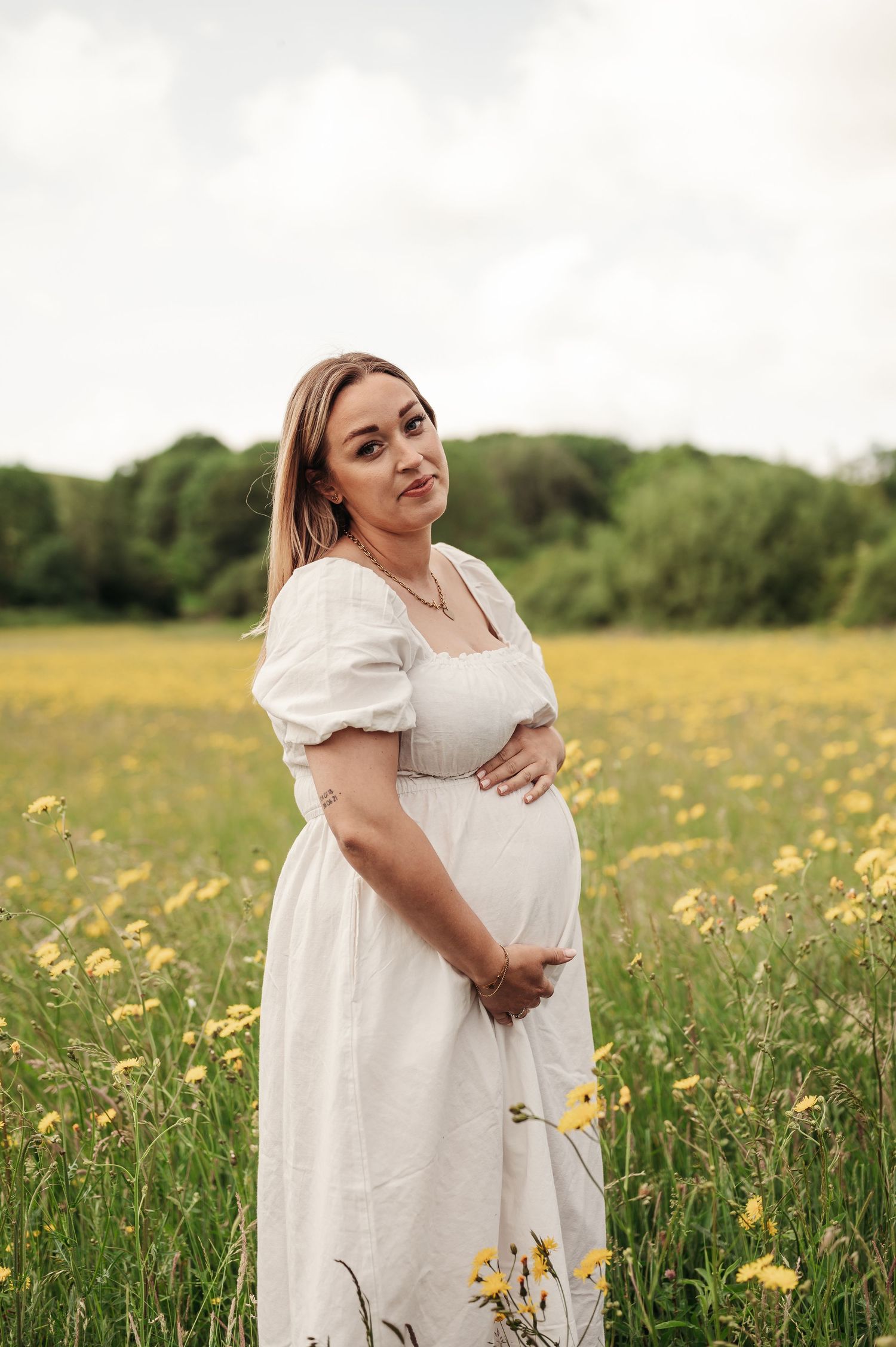 Someone in a flowing white dress stands in a field of yellow wildflowers during a scenic outdoor maternity photo session.
