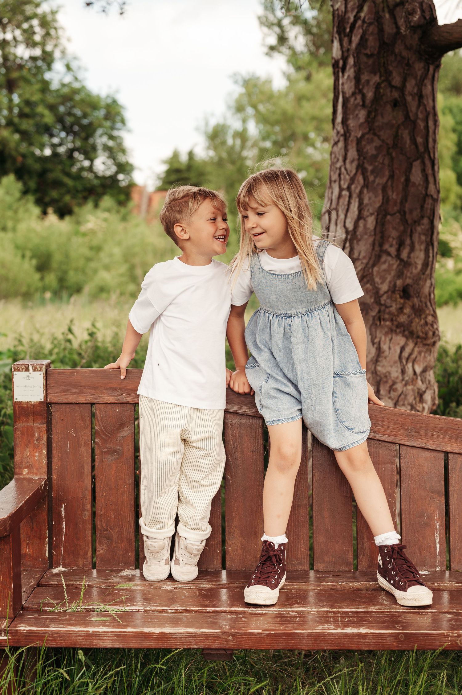 Two children share a playful moment together while standing on a brown wooden deck by a large pine tree.