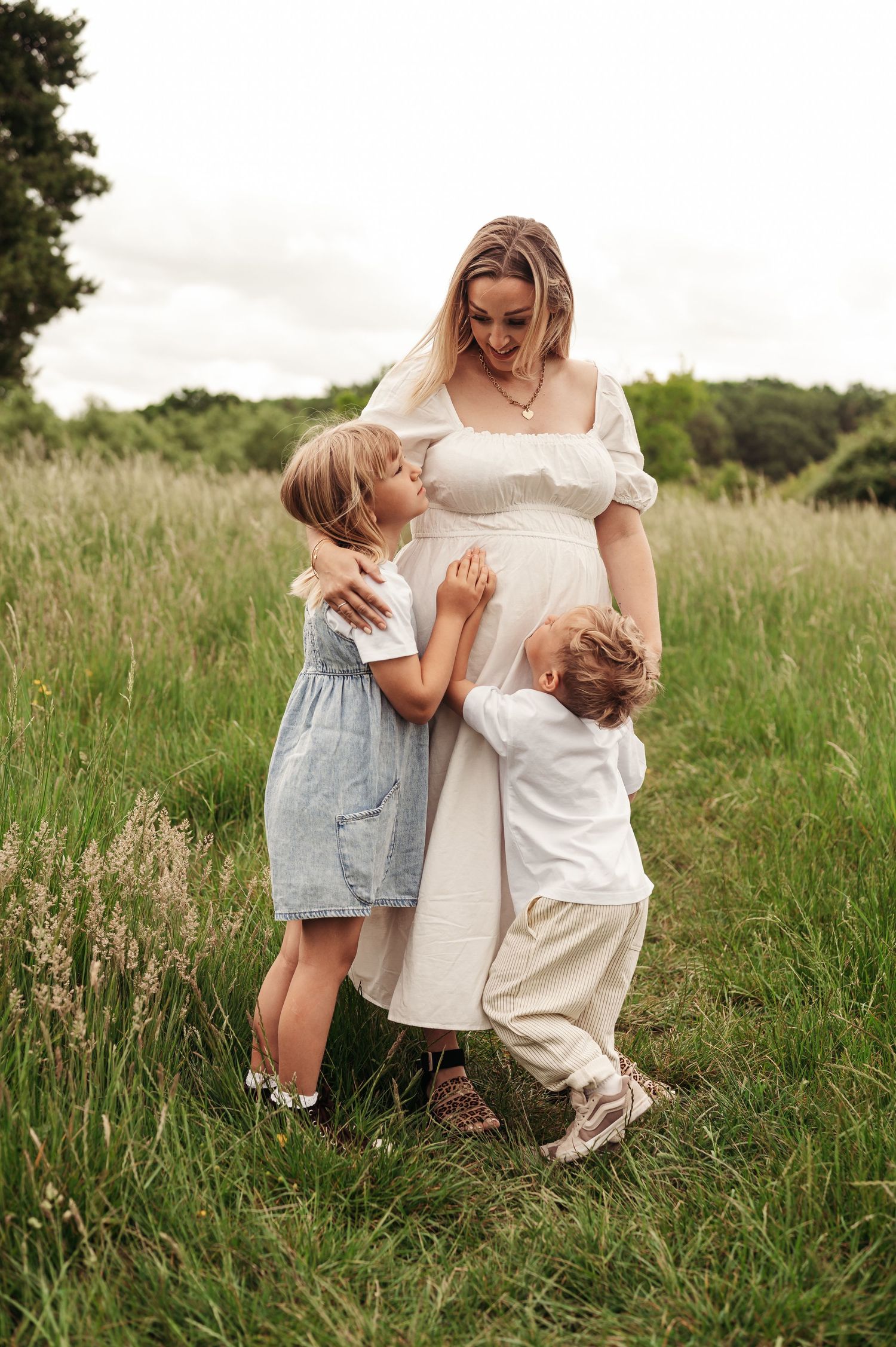 A joyful moment in a summer meadow as children embrace a pregnant woman wearing a white dress.