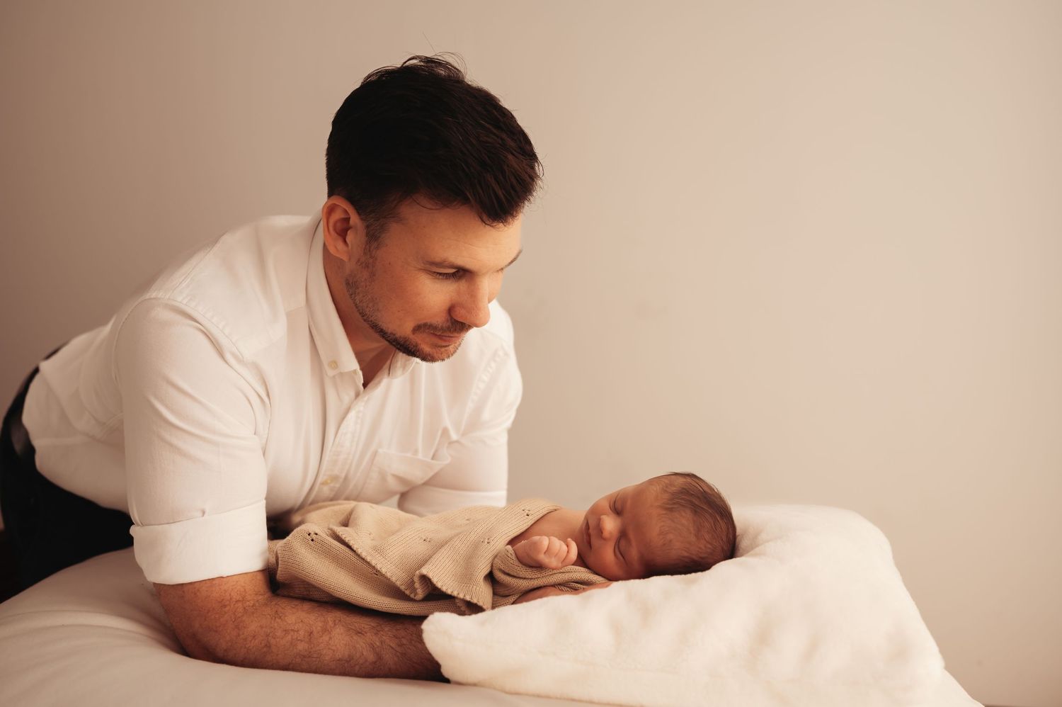 Parent gazing lovingly at sleeping newborn wrapped in beige blanket on white bed.