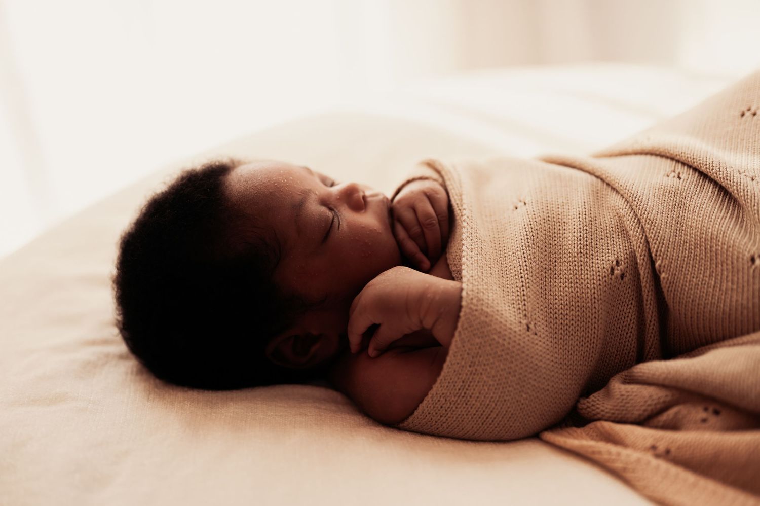 Newborn baby swaddled in a soft beige knit blanket sleeping peacefully on a light-colored bed.