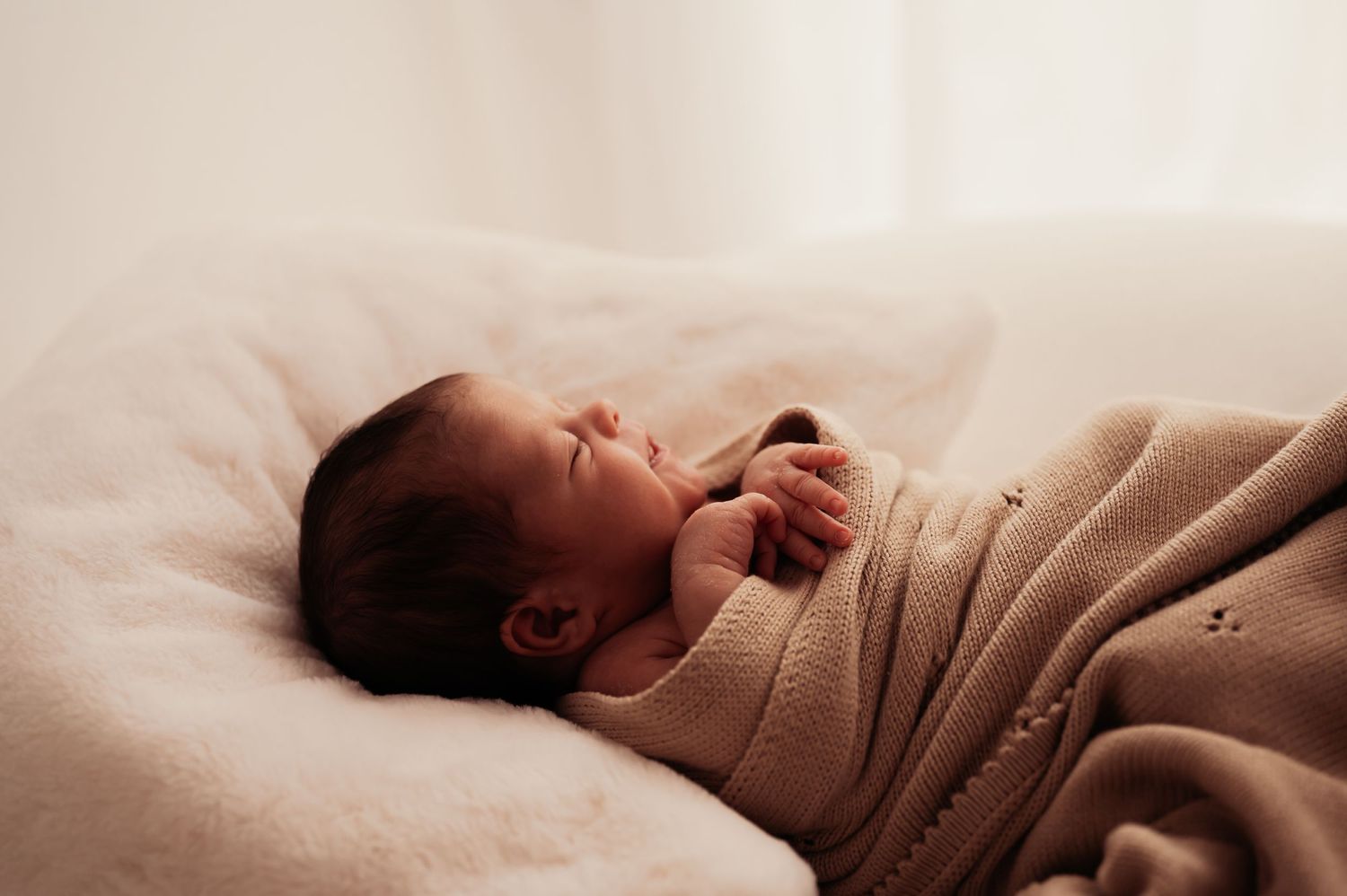Newborn baby sleeping peacefully while wrapped in tan knit blanket on white bedding.