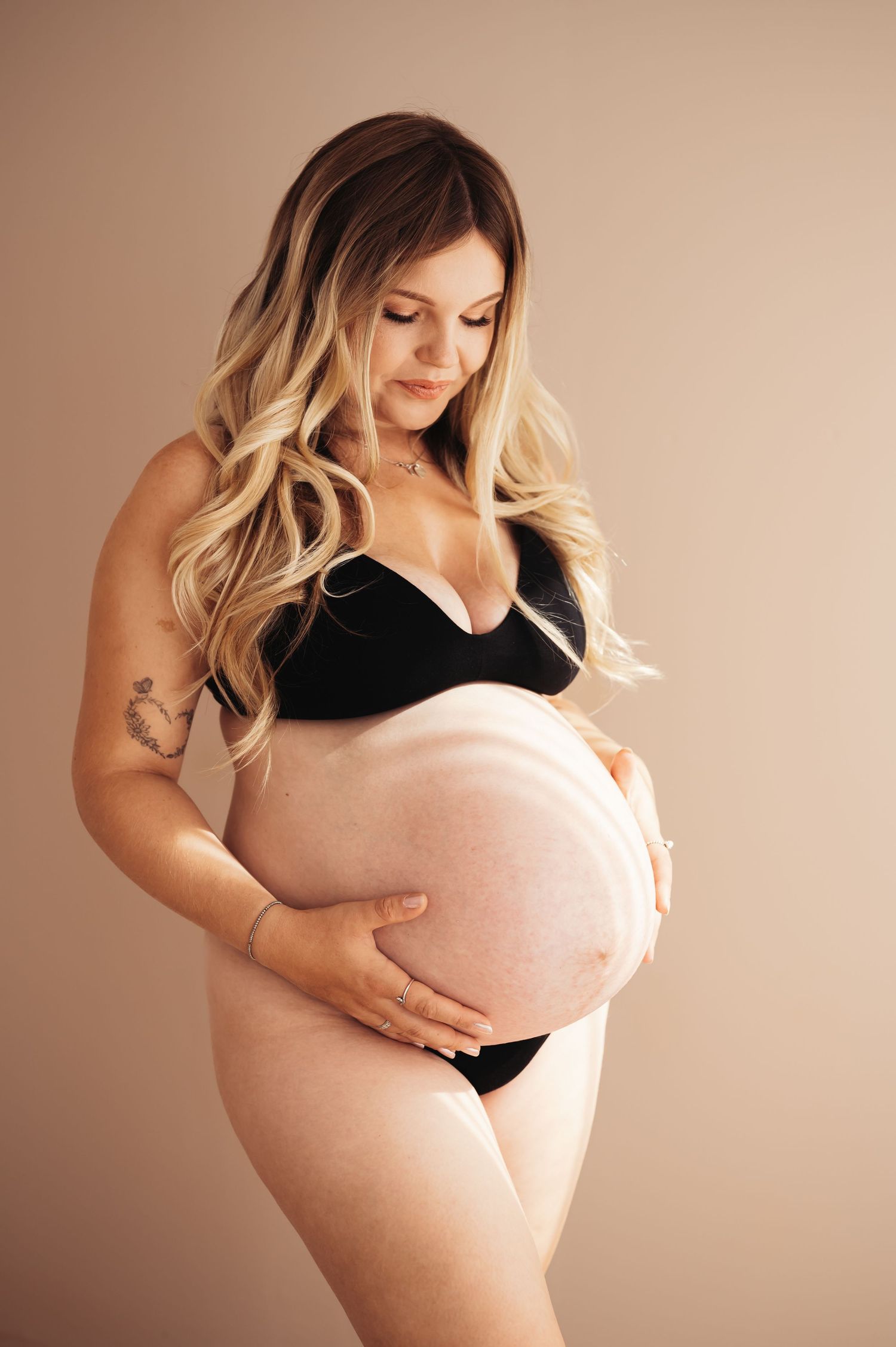 Pregnant woman in black lingerie cradling her baby bump, smiling down with long blonde hair in a studio setting.
