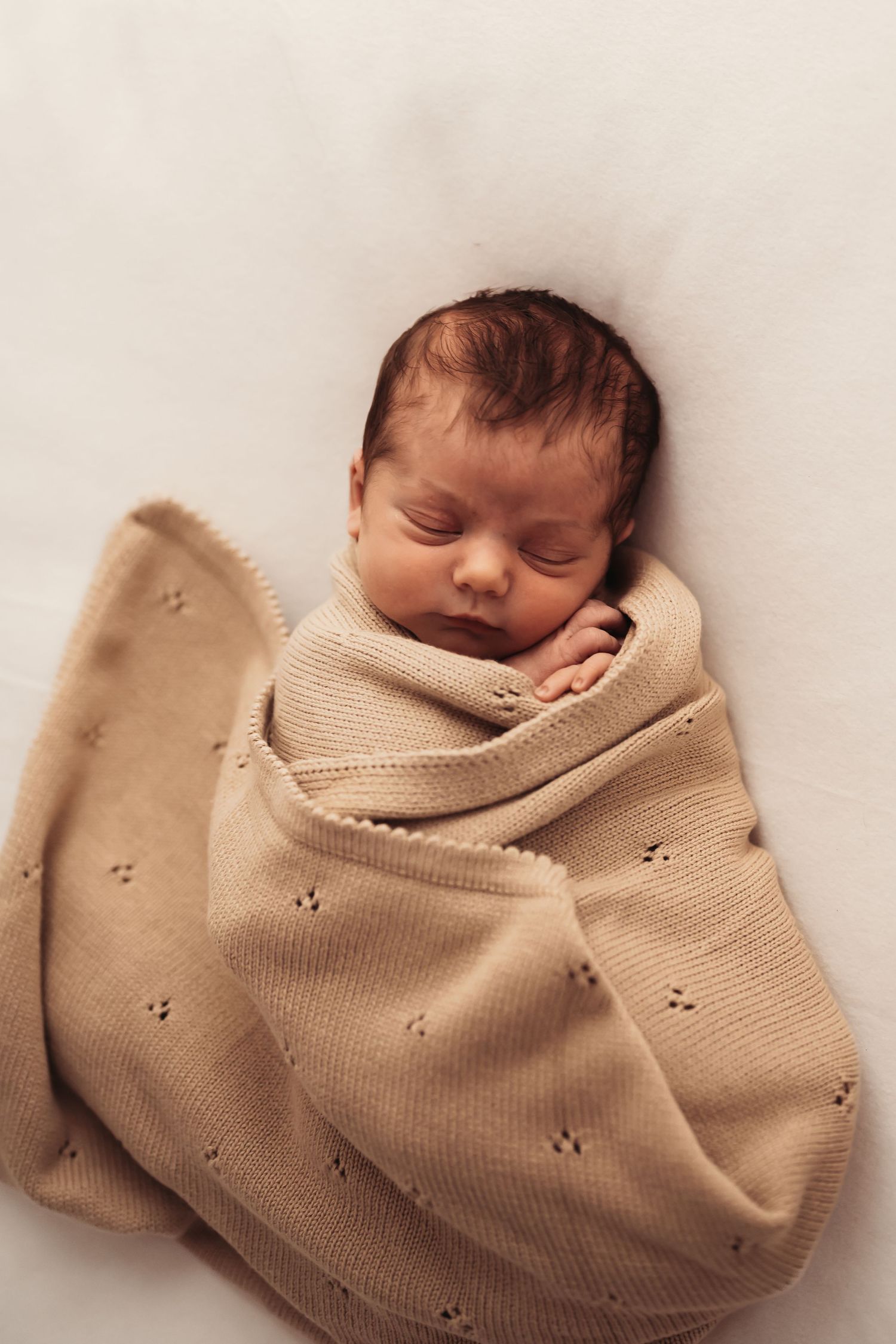 Sleeping newborn wrapped in beige knit blanket during professional photo session.