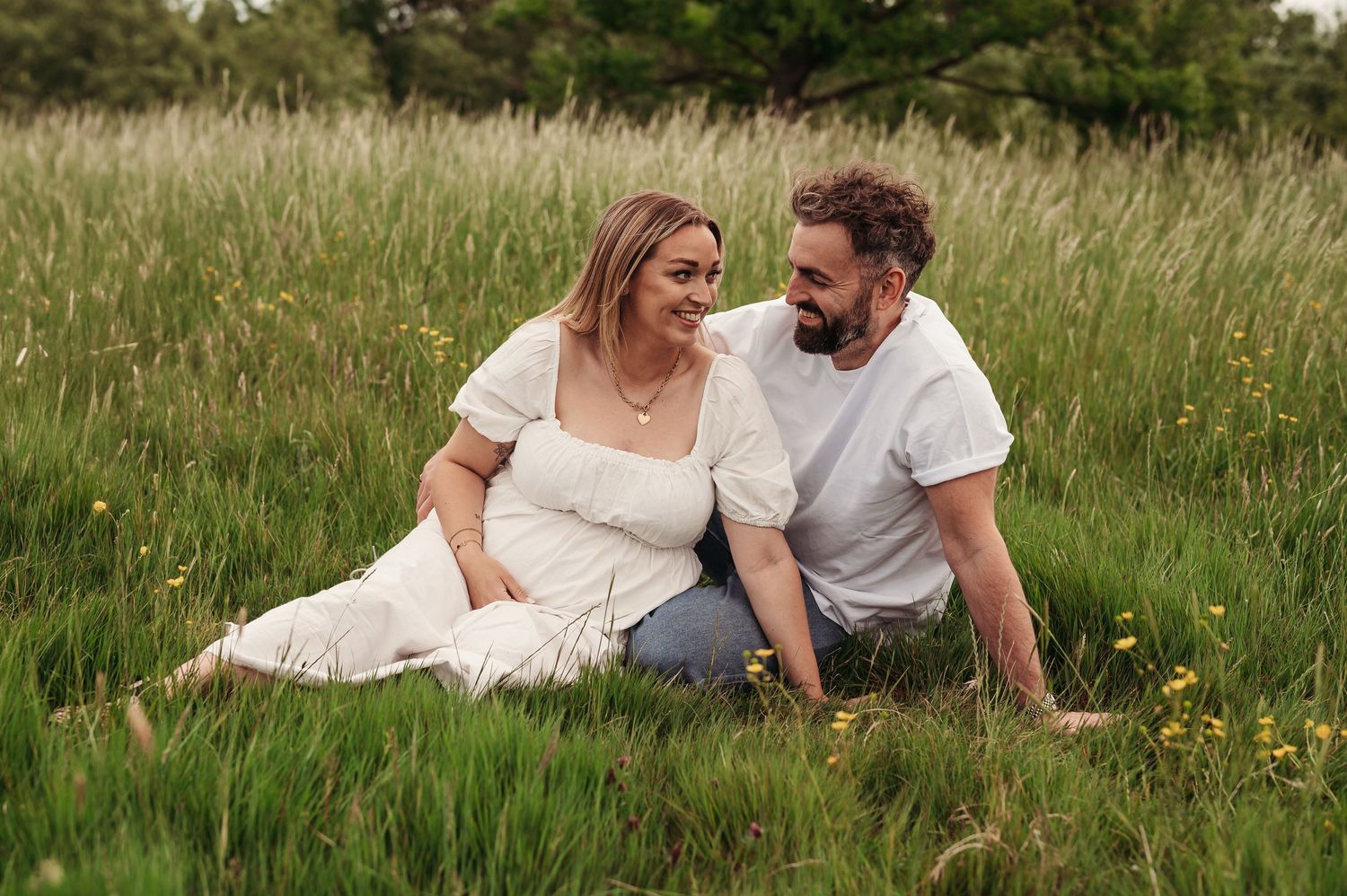 A couple sits together in a sunlit meadow with tall grass and wildflowers on a summer evening.