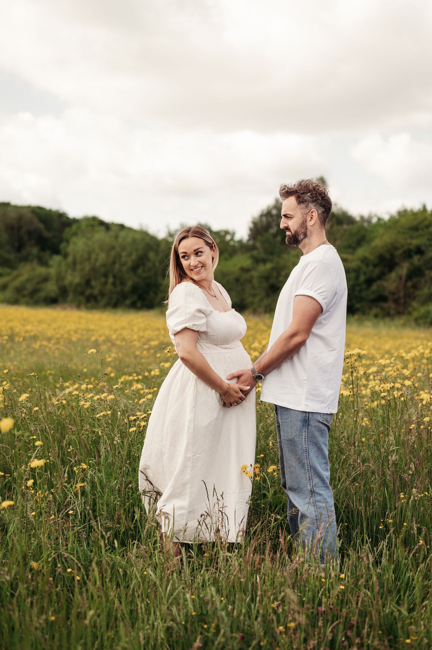 A couple stands together holding hands in a sunny meadow filled with yellow wildflowers and tall grass.
