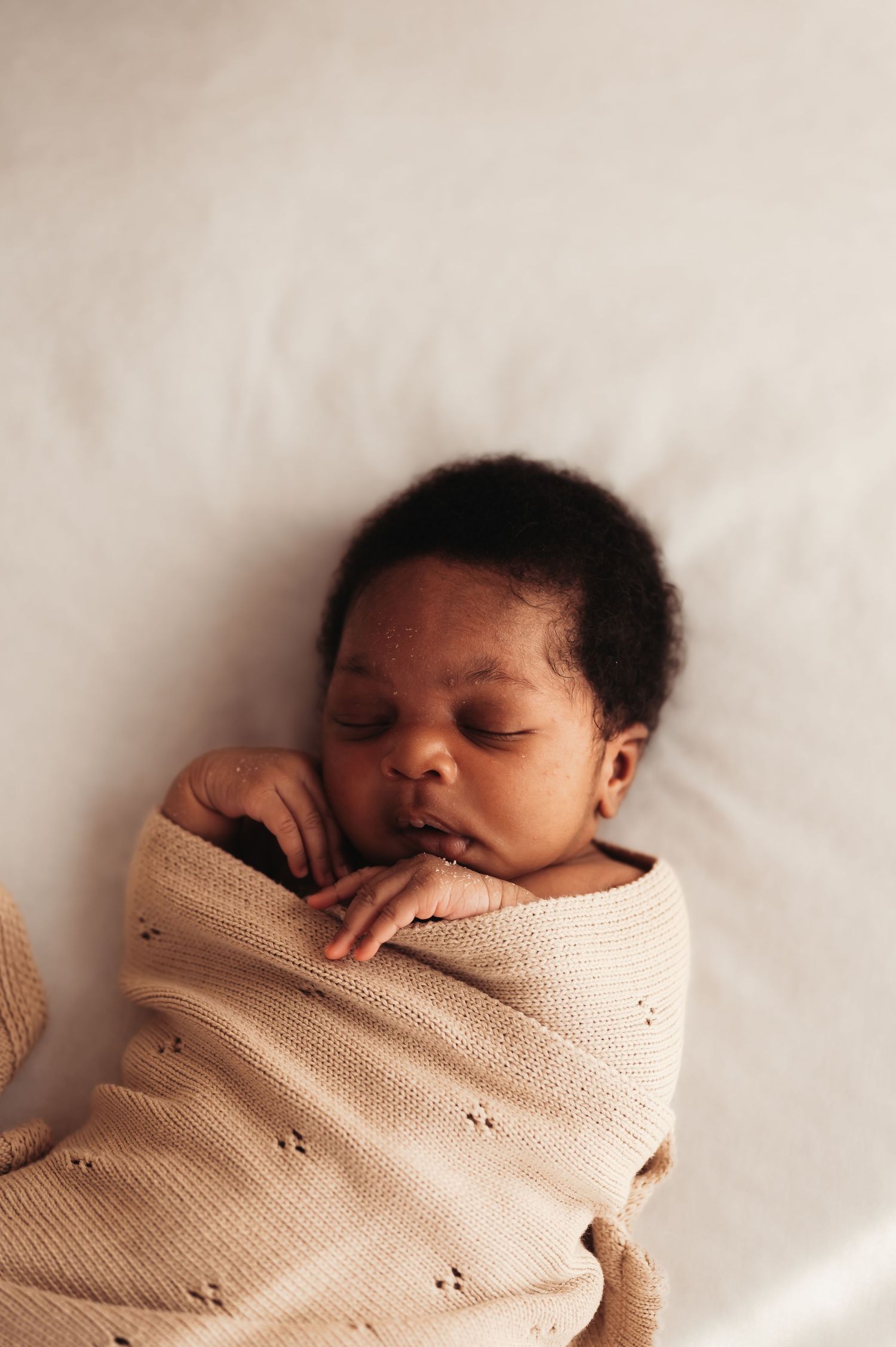 Peaceful newborn baby swaddled in a soft beige knit blanket, sleeping on a white surface.